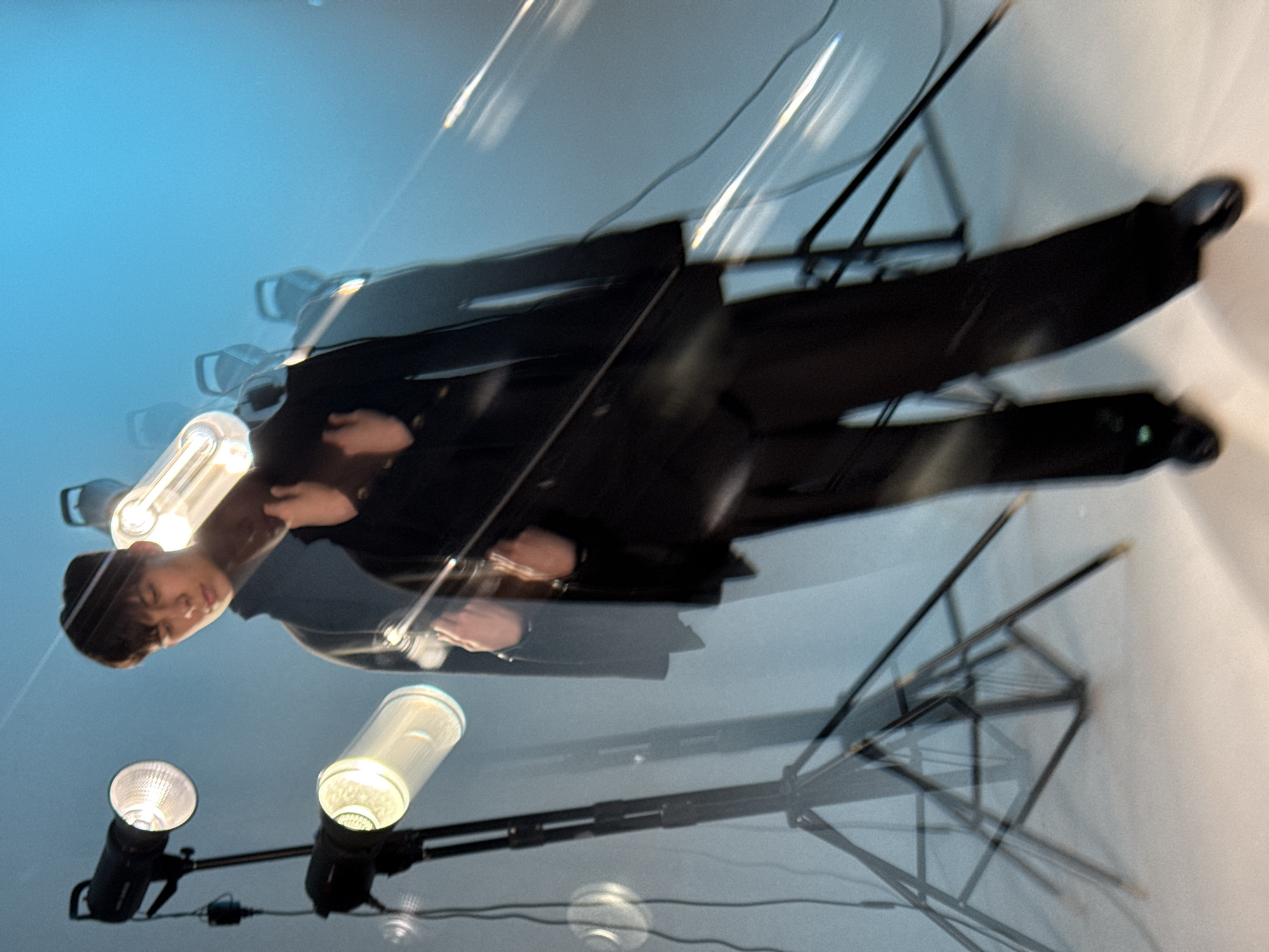 The image shows a reflection of a person in a black suit, with studio lighting equipment, in a likely indoor setting. There are visible light stands with studio lights, including a strobe. The reflection suggests a glossy surface, possibly a polished floor. The environment is a studio or similar space prepared for a photoshoot. There is no information that suggests the location is the city of Chaikovsky, Russia.