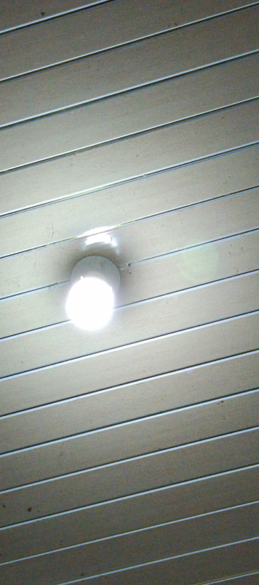 A ceiling light fixture is visible in the center of the image, mounted on a surface composed of horizontal, parallel slats. The light is illuminated and emits a bright glow, casting a soft reflection on the surrounding slats. The slats appear to be made of a light-colored material, possibly wood or plastic, with thin, dark lines separating them. The overall scene suggests an indoor environment, likely a room or hallway with a paneled ceiling. There is no information available to confirm the location as Pashiebeutong, Indonesia.