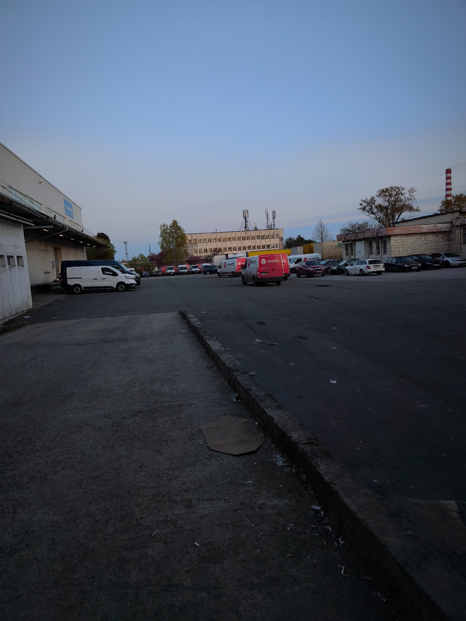 This image presents an industrial or commercial yard in Burgas, Bulgaria, captured during either dawn or dusk, as suggested by the clear, pale blue sky and the muted, shadowed lighting across the scene.
On the left, a large, white warehouse building features loading docks, and a blue sign with the partially visible text "TIMBEREX" is affixed to its facade. Several white commercial vans are parked alongside this building. The foreground and midground are covered by an asphalt parking lot, where numerous vehicles are stationed. Prominently featured is a red delivery van marked with the "Speedy" logo on its side and rear, with another "Speedy" van visible further back. A variety of other cars and vans are also parked throughout the lot.
In the background, additional industrial buildings with multiple windows and rooftop communication antennas can be seen. A distinctive red and white striped chimney rises on the far right, acting as an industrial landmark. Scattered trees, some with autumnal foliage, punctuate the landscape. The asphalt ground features a concrete curb along the right, with some debris, including cardboard and what appear to be cigarette butts.
The scene conveys the functional atmosphere of a logistics or commercial hub. While no specific event is actively taking place and no people are visible, the setup strongly implies ongoing operations involving delivery, storage, or industrial activities within this Bulgarian setting.