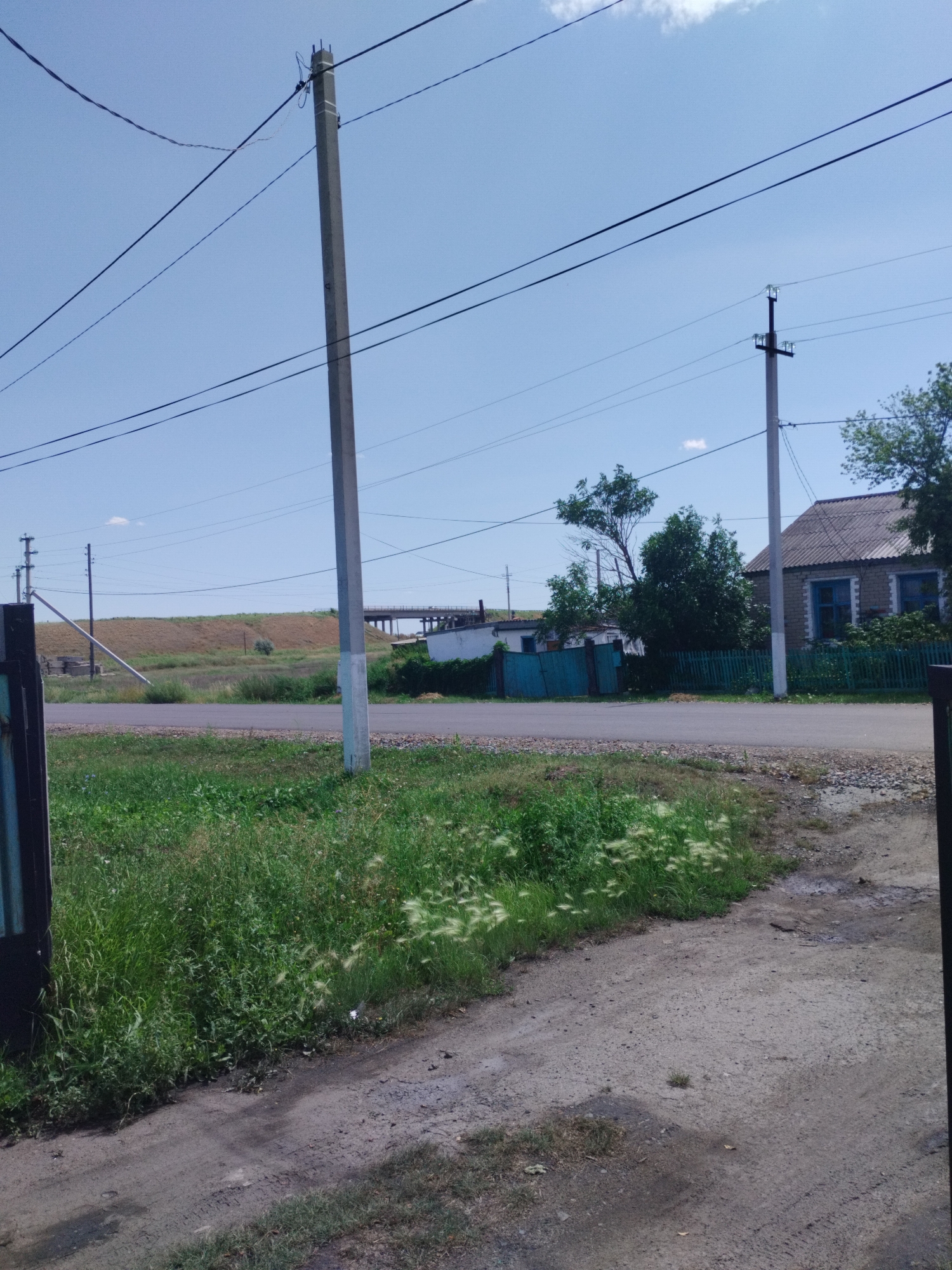 The image shows a rural or semi-rural street scene with a clear sky and some clouds. In the foreground, a dirt road leads into the scene from the right, bordered by a patch of green grass and some taller weeds with feathery seed heads. To the left, a dark metal gate post is visible. A paved road runs horizontally across the middle ground.
Along the paved road, there are several utility poles with wires. The closest utility pole on the right is a concrete pole with supporting insulators and wires. Further down the road, another utility pole is visible. On the right side of the paved road, a house with a grey, textured roof and blue-trimmed windows is situated behind a green wooden fence. Adjacent to this house, there is a smaller white building with a blue-green gate. Trees and bushes are present in this area.
In the background, a grassy embankment rises towards a structure that appears to be a bridge or an overpass. Another concrete utility pole is located near the base of the embankment on the left side of the paved road. The overall impression is of a quiet, possibly sparsely populated area. The location is identified as Sarykol, Kazakhstan.