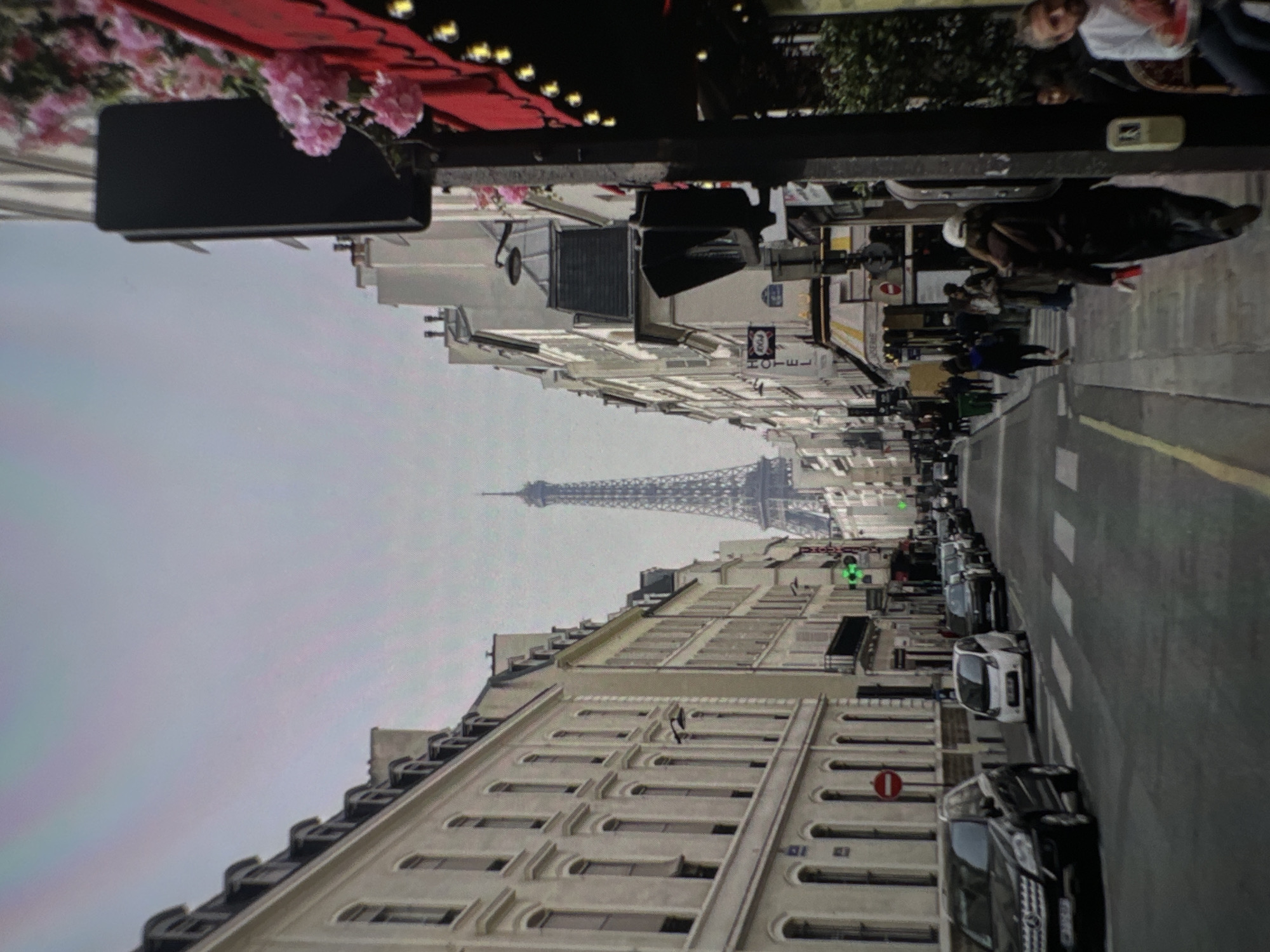 The image shows a street view with the Eiffel Tower in the distance. The street is lined with buildings on both sides. There are cars parked along the side of the street and people walking on the sidewalk. There is a red awning and flowers visible in the foreground. This is Paris, France, not Chișinău, Moldova.