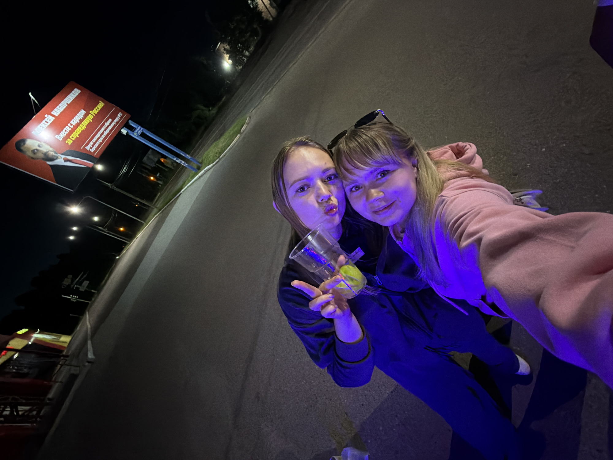 In the city of Chaikovsky, Russia, at night, two young women are taking a selfie on a street. A billboard featuring a man in a suit and tie is visible in the background. One woman is holding a plastic cup with a lime slice. Both women are illuminated by purple light. The overall scene appears to be a casual, nighttime gathering.