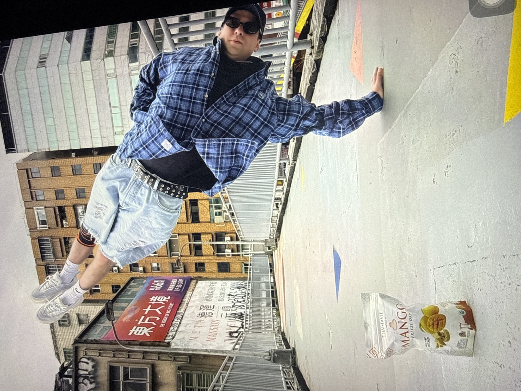 A young person is performing a handstand on a pedestrian overpass in Taipei, Taiwan. The person is wearing a blue plaid shirt, ripped denim shorts, white socks, and white sneakers. They are also wearing sunglasses and a black belt. In the foreground, a bag of freeze-dried mangoes is lying on the ground. In the background, there are several buildings, including a tall, modern skyscraper and older, more dilapidated structures. The overpass itself is made of concrete and has a metal railing on either side. There are colorful geometric shapes painted on the overpass surface. The sky is overcast.