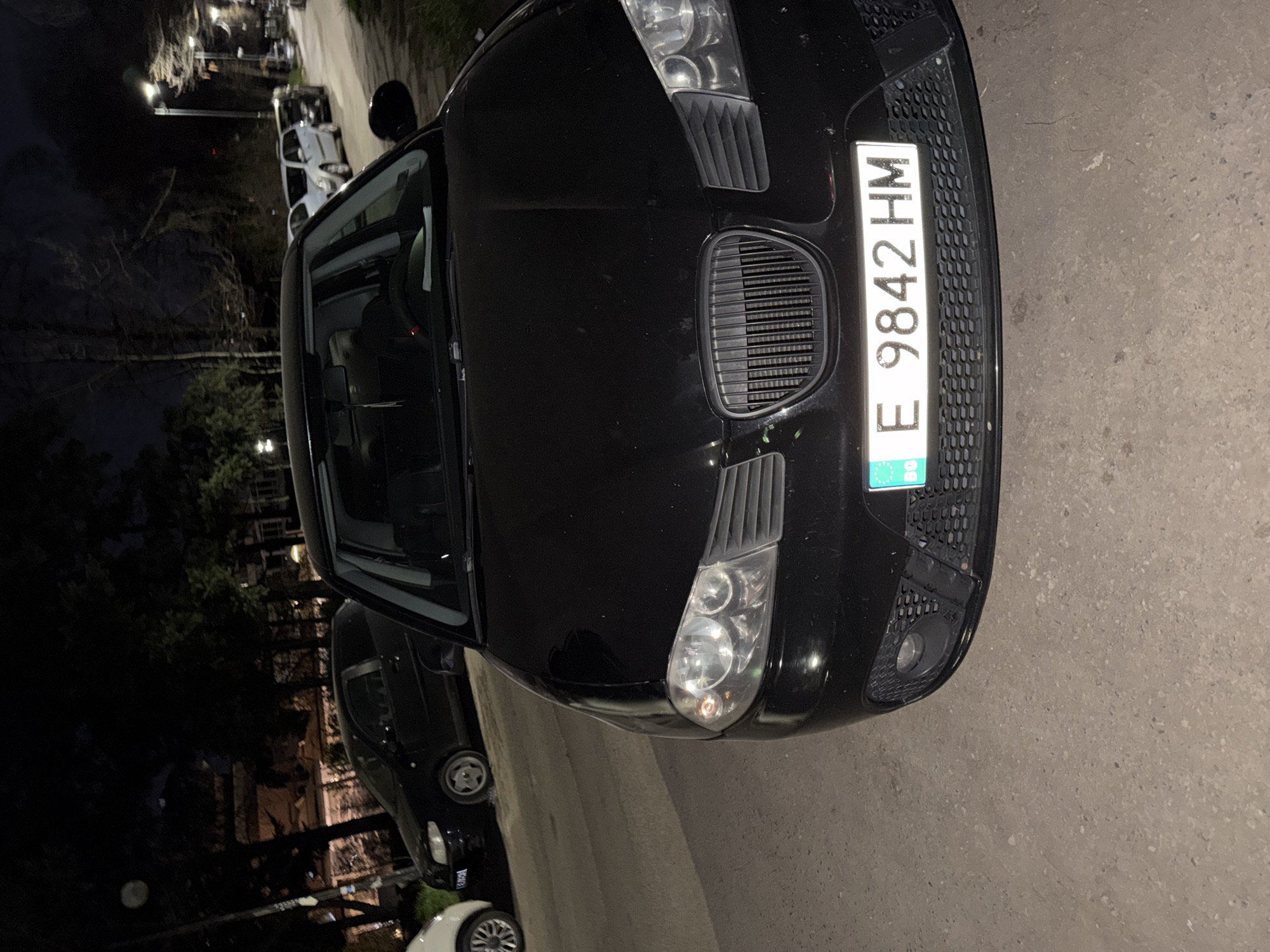 The image shows a black car parked on a street. The car's license plate reads "E 9842 HM" and displays the "BG" country code, indicating it is registered in Bulgaria. The vehicle is parked at night on a street in what appears to be the city of Sofia, Bulgaria. Other cars are visible in the background. The lighting is dim, and the surrounding environment includes trees and streetlights.