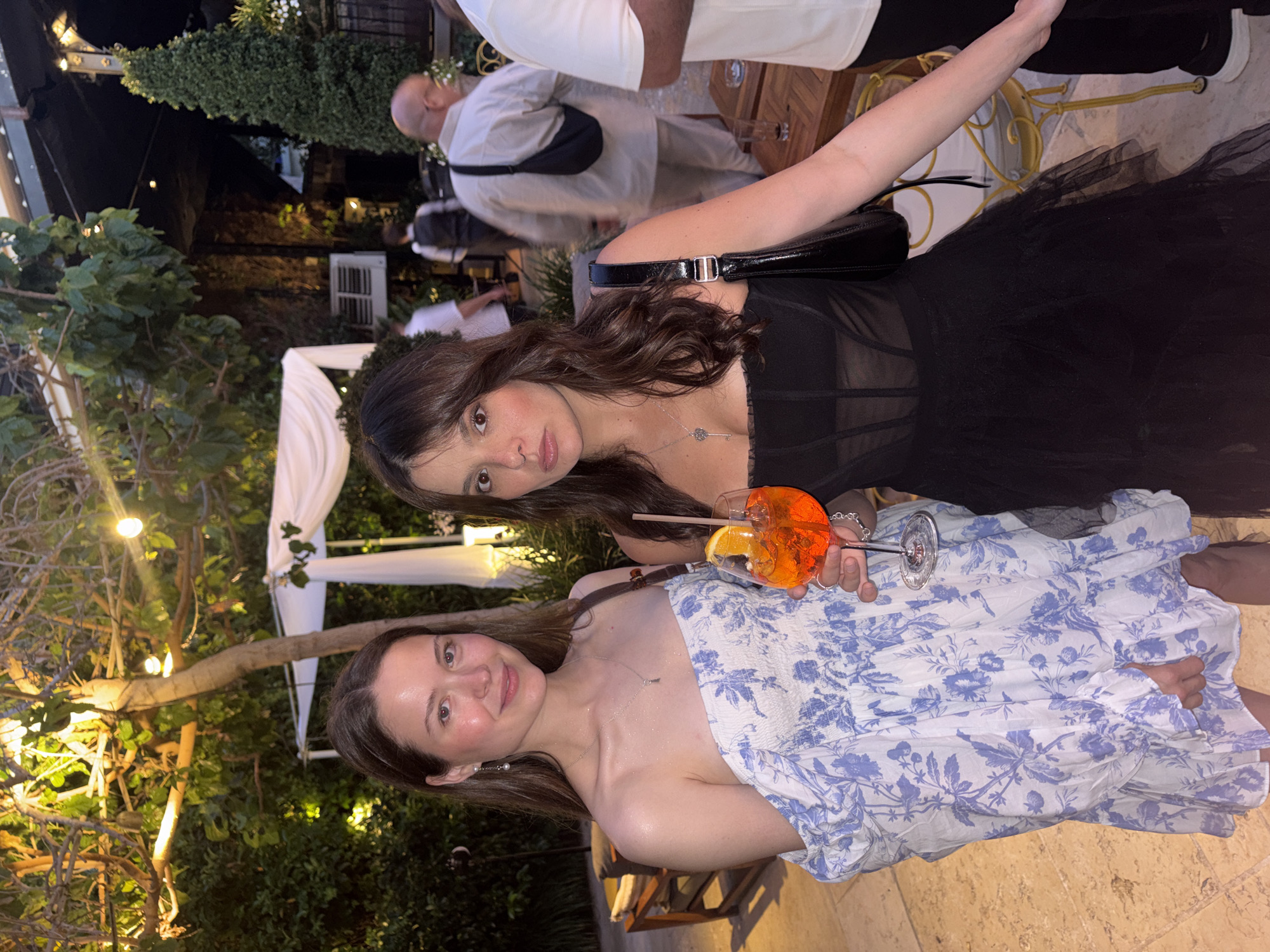 Two young women pose for a photograph outdoors. One woman wears a strapless, white dress with a blue floral print. She holds a cocktail in a stemmed glass with a straw and an orange slice garnish. The second woman wears a black dress with a tulle skirt, and a black purse over her shoulder. They are in a social setting with other people visible in the background. The environment includes trees, plants, and a white canopy structure illuminated with soft lighting. The location is assumed to be Tel-Aviv, Israel, based on contextual information.