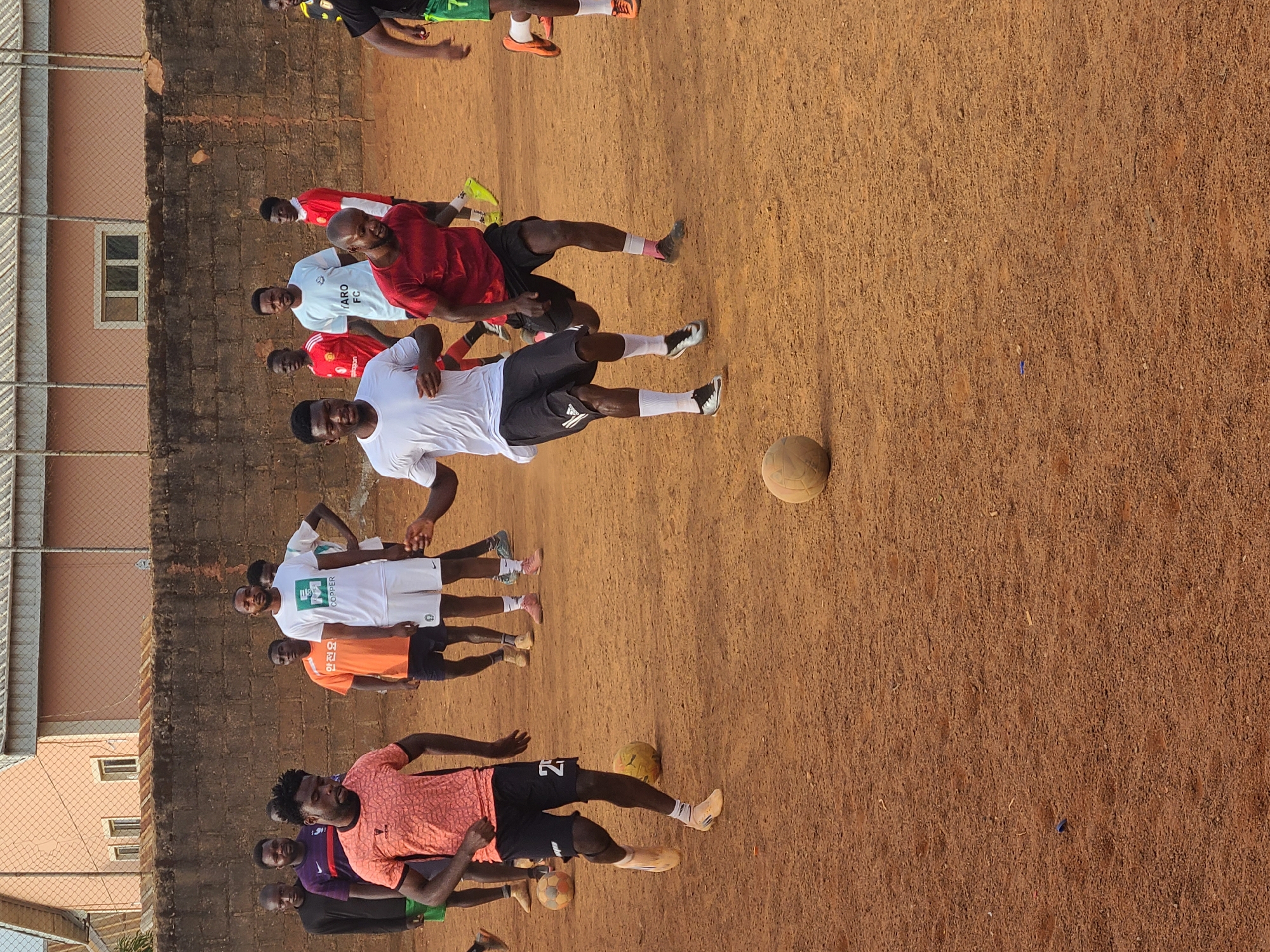 The image depicts a group of approximately ten men playing soccer on a dirt field. The men are wearing various athletic shirts and shorts. Several soccer balls are visible on the ground. A brick wall runs along one side of the field, and a building with a window and a chain-link fence are in the background. The location appears to be in a city, Unknown, Unknown.