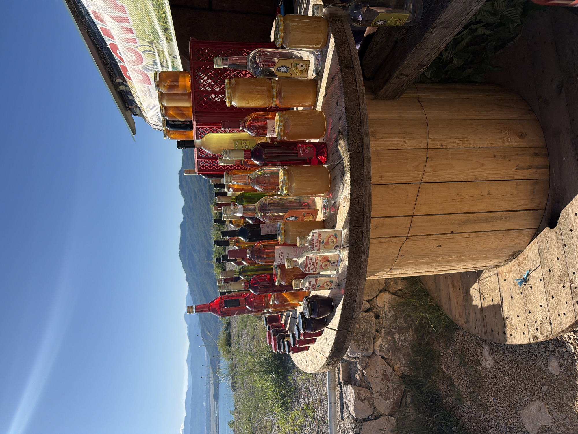 In Ponikvica, Montenegro, an outdoor market stall displays an assortment of bottled beverages and jars of preserves. The stall is set up on a large, wooden spool, with a stone wall and dry vegetation in the foreground. In the background, a body of water and rolling, green hills under a clear blue sky suggest a rural or coastal setting. Sunlight casts long shadows, indicating it is likely late afternoon.
The table is laden with numerous bottles of various shapes and sizes, containing liquids of different colors, from clear to deep red. Many of these appear to be alcoholic beverages, such as wine and spirits, judging by their labels and bottle designs. Several small glass jars filled with golden-colored preserves, likely honey or fruit preserves, are also present. Some jars have labels with images of fruit, possibly indicating the flavor. A red plastic crate is visible behind some of the bottles, holding more jars.
The overall impression is that of a local vendor selling homemade or artisanal products in a picturesque, natural setting. No people are visible in the frame. The scene is static and peaceful, with a focus on the products and the surrounding landscape.