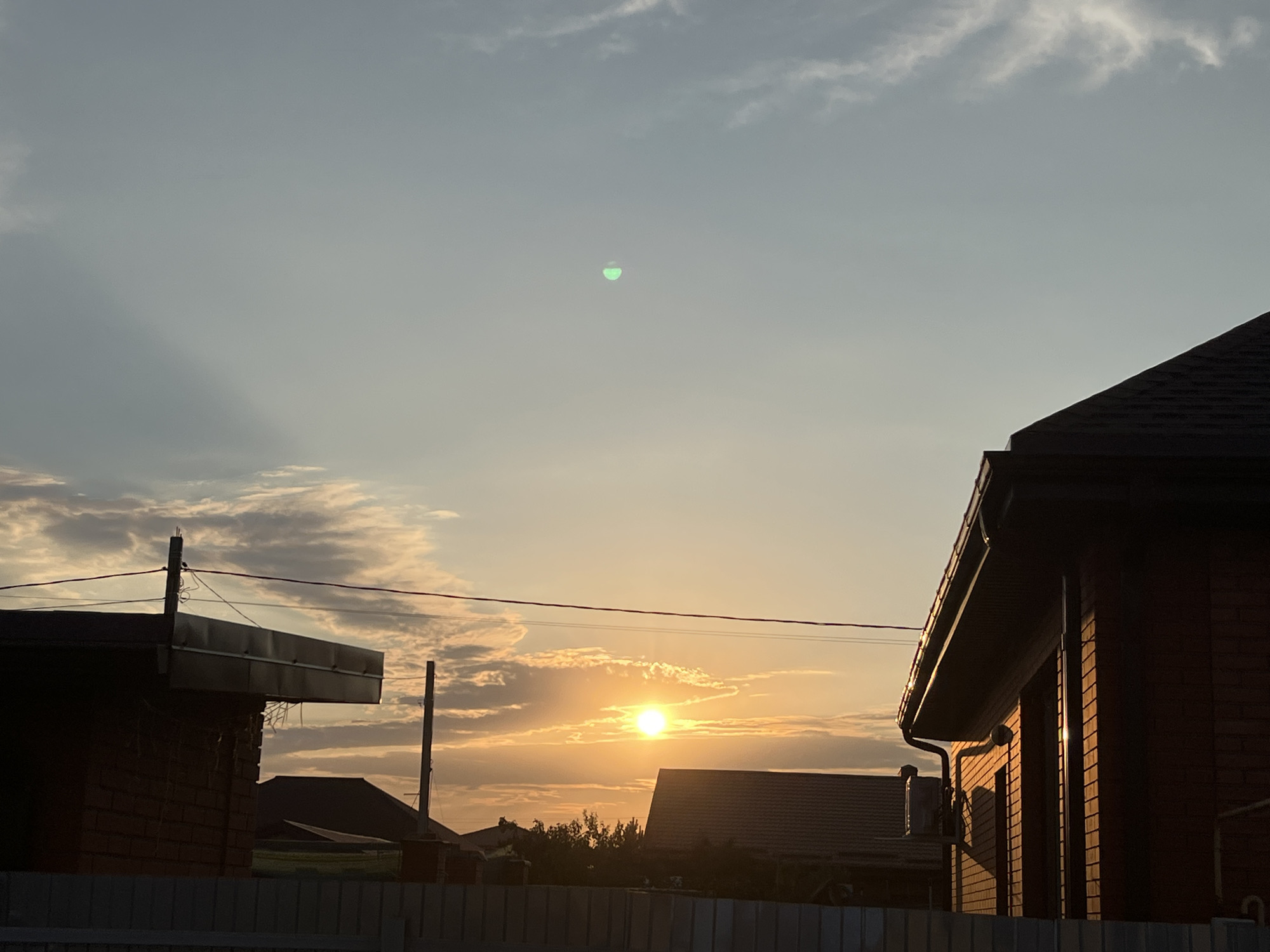 The image captures a sunset over a residential area, likely in Taganrog City District, Russia. In the foreground, a light-colored fence runs horizontally across the bottom of the frame. Behind the fence, brick buildings with dark roofs are visible. The building on the left has a flat, dark-colored roof overhang. The building on the right has a sloped, dark roof and brick facade with vertical lines indicating shadows. In the middle ground, several other houses with dark roofs are silhouetted against the sky. Trees are visible between some of the buildings. The sky is a gradient of colors, transitioning from a pale blue at the top to warmer oranges and yellows near the horizon. The sun, a bright orange orb, is partially obscured by thin clouds. Faint shadows stretch across the sky from the left. A small, bright green, oval-shaped object is present in the upper portion of the sky. Utility poles and wires are visible, suggesting an active residential area. The scene appears to be devoid of any human presence.