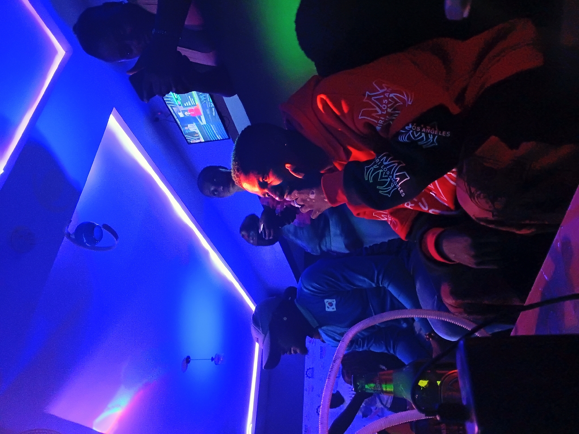 The image depicts an indoor scene, likely a lounge or club, with vibrant blue and green neon lighting illuminating the space. In the foreground, a person wearing an orange hooded sweatshirt with white graphic designs is seated, looking to their right. To their left, another individual in a blue long-sleeved shirt and a baseball cap is also seated, facing left. Both individuals appear to be in conversation or engaged with their surroundings. In the background, other people are visible, some holding phones. A television screen mounted on the wall displays an image, and a shisha pipe with hoses is present on a table in the lower left quadrant of the image. The overall atmosphere suggests a social gathering. The presence of the television screen, shisha pipe, and the general ambiance indicate it could be a popular spot for entertainment and socializing in Jos, Nigeria.