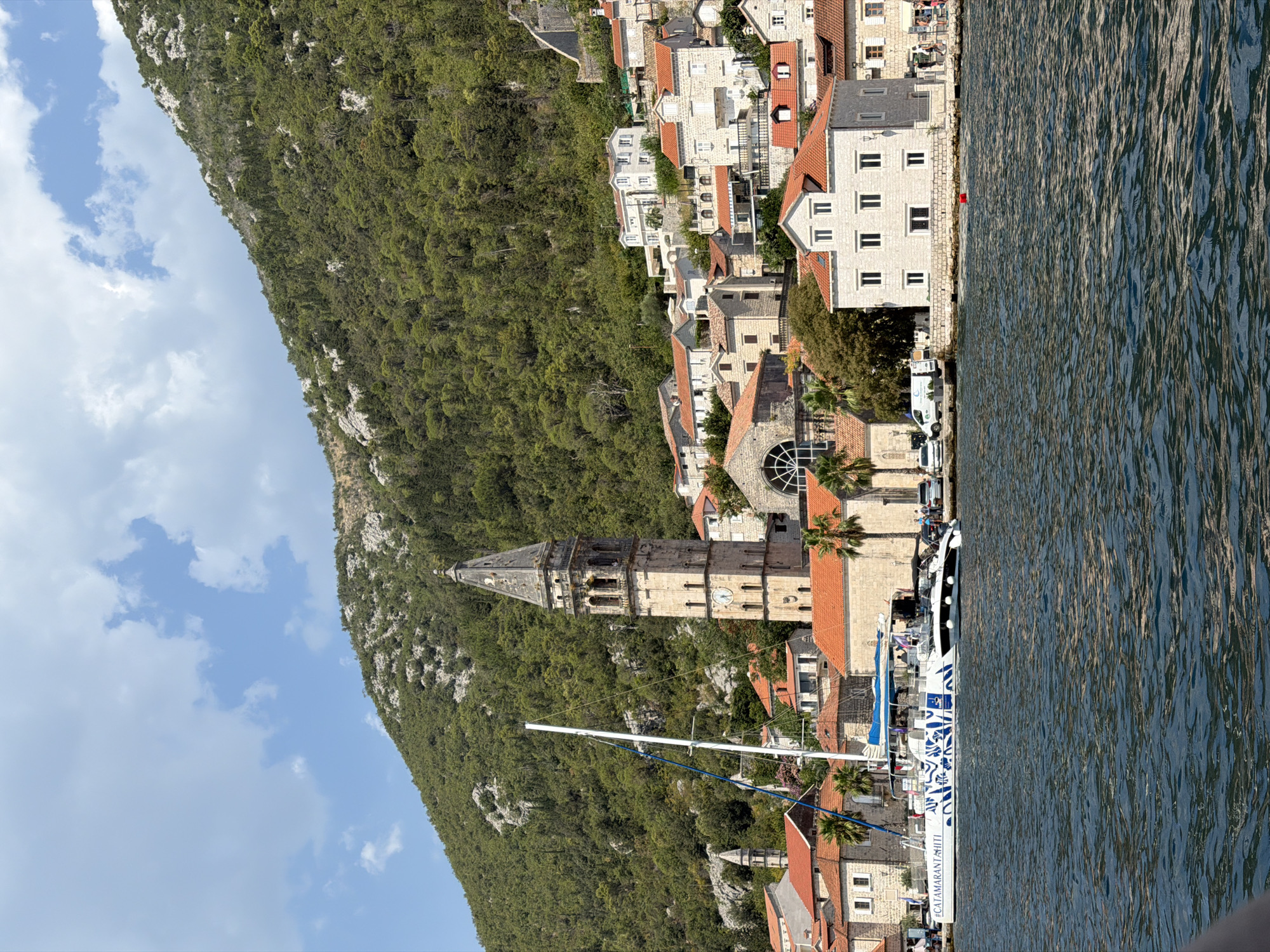 The image depicts a picturesque coastal scene in Perast, Montenegro. In the foreground, the calm, dark blue water of the bay is visible, with gentle ripples reflecting the sunlight. A white catamaran, adorned with blue graphic designs and the hashtag "#CATAMARANTAHITI" on its side, is moored near the shore. Several people are visible on the deck of the catamaran.
Behind the catamaran, a stone promenade lines the waterfront, with a few palm trees and dense green vegetation interspersed between buildings. The town of Perast rises up a steep hillside, characterized by its traditional stone buildings with red-tiled roofs. Dominating the skyline is the tall, weathered bell tower of a church, featuring a clock face and arched openings. Other church towers and structures are also visible on the slope. The hillside behind the town is covered in lush, green trees and rocky outcrops. The sky above is a clear blue, with a few scattered white clouds. The scene conveys a sense of tranquility and historical charm, typical of coastal Mediterranean towns.