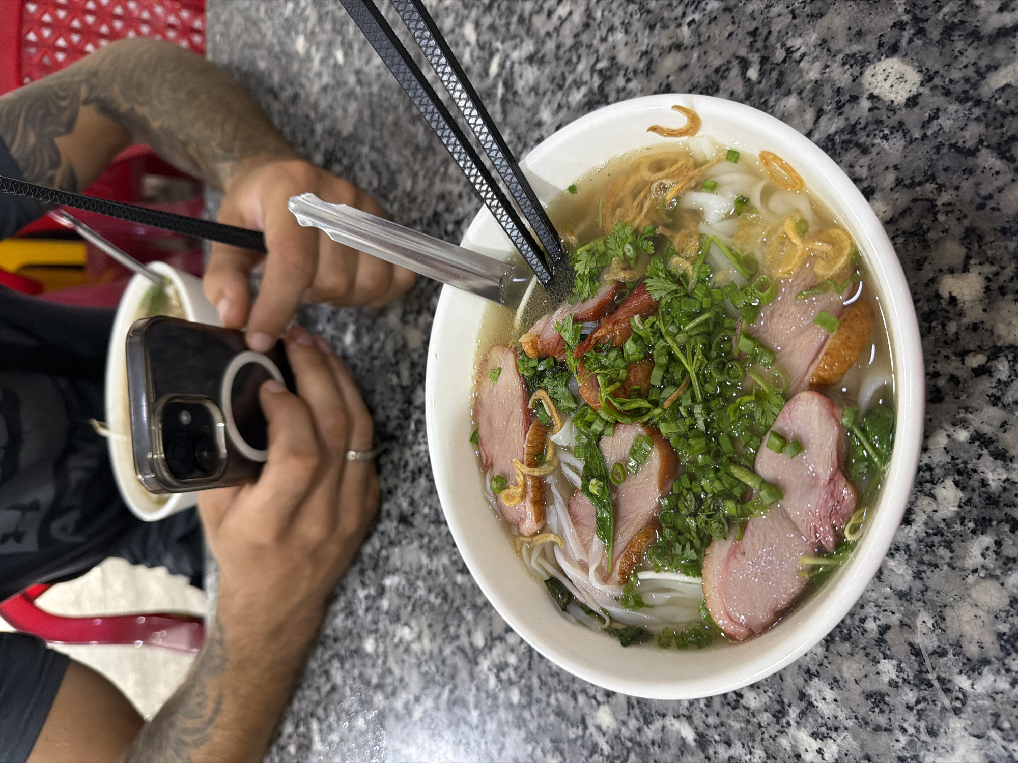 The image presents an overhead view of a person capturing a meal with a smartphone at a grey speckled granite table. The main subject is a large white bowl of Vietnamese noodle soup, prominently displayed in the center-right of the frame. The soup contains wide flat rice noodles, slices of roasted meat (appearing to be duck or char siu pork), a generous topping of chopped green onions and cilantro, and crispy fried shallots in a clear broth.
A pair of black, patterned chopsticks rest across the rim of the bowl, with a silver spoon submerged in the broth. The person's forearms, visible on the left side of the image, are heavily tattooed. Their left hand holds a black smartphone, equipped with a clear case and a circular accessory (likely a MagSafe ring), angled to photograph the noodle dish. A ring is visible on a finger of the person's right hand, which is also holding the phone. A smaller, partially visible white bowl is to the left of the main dish.
The setting appears to be an indoor dining area, such as a restaurant or eatery, suggested by the table surface and the nature of the meal. A blurry red object, possibly a chair or part of the restaurant decor, is visible in the background. The lighting is soft and even, typical of an indoor environment. The scene captures the modern practice of documenting food before eating. No discernible text is visible within the image.