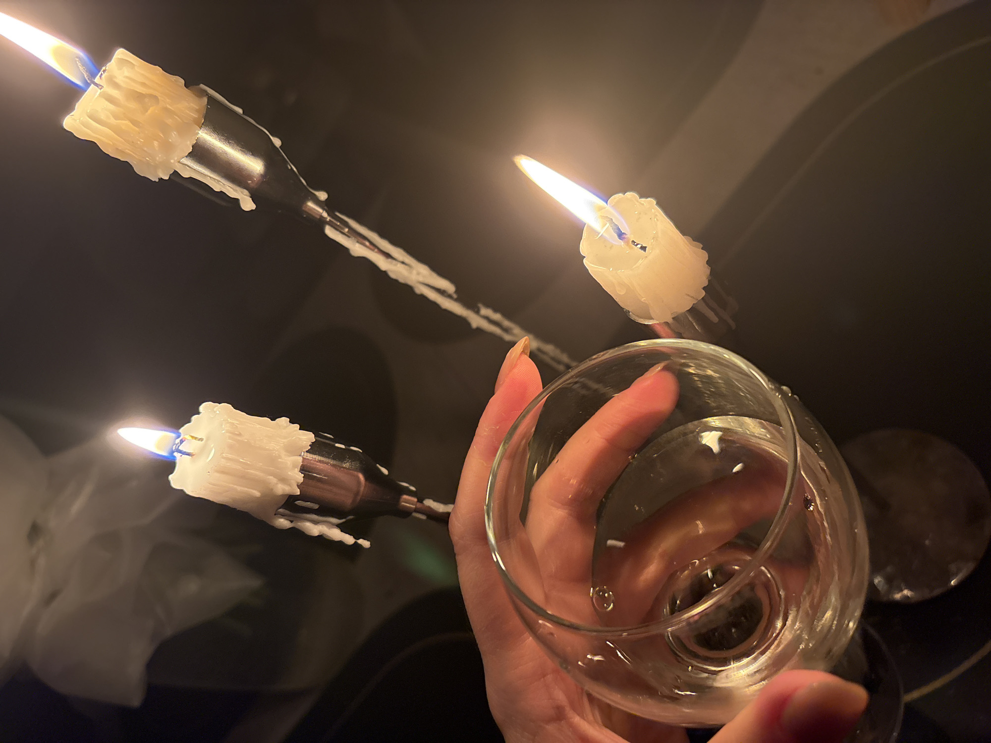 A low-angle shot shows a hand holding a clear wine glass with liquid, surrounded by three makeshift candle holders with lit candles. The candle holders appear to be made from metallic objects and feature partially melted white candles. The background is dark, with surfaces exhibiting reflective qualities. The location is assumed to be in the city of Chaikovsky, Russia. There is one hand visible.