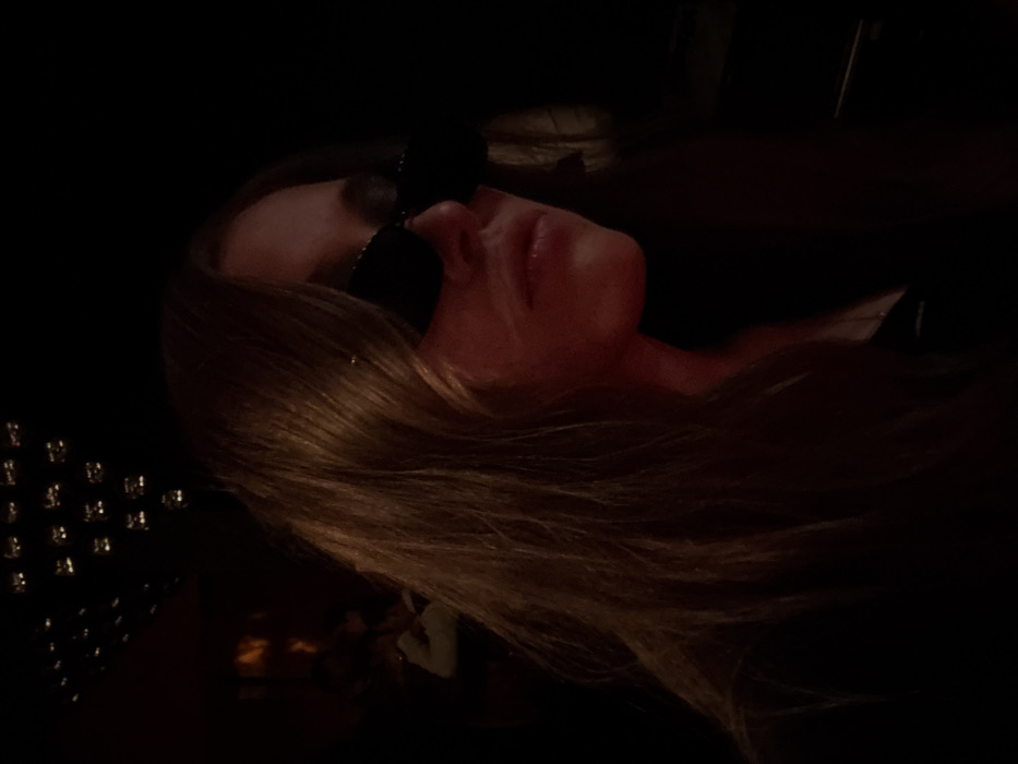 A person wearing sunglasses lies in a dimly lit setting. Light reflects on their skin, lips, and blonde hair, which flows across the lower half of the frame. A dark, textured surface, adorned with reflective dots, is visible in the lower left. The background appears dark and undefined. The individual's neck and a hint of a shoulder are visible against the black backdrop.
