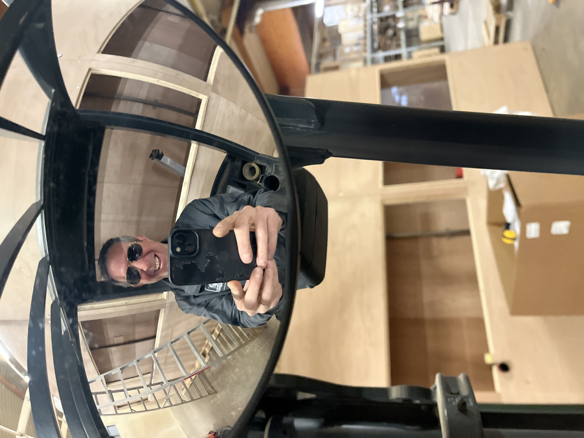 A man with sunglasses smiles at his phone while holding it up to a helicopter's cockpit mirror. The reflection shows the man standing inside the unfinished cockpit which has wooden frames for windows. A ladder leans against the inside wall. Through the helicopter's rotor hub, the image shows a large open warehouse space with shelving, boxes, and unfinished wooden cabinets. The location is likely within an aviation-related business in Leudelange, Luxembourg, given the presence of the helicopter and manufacturing elements.