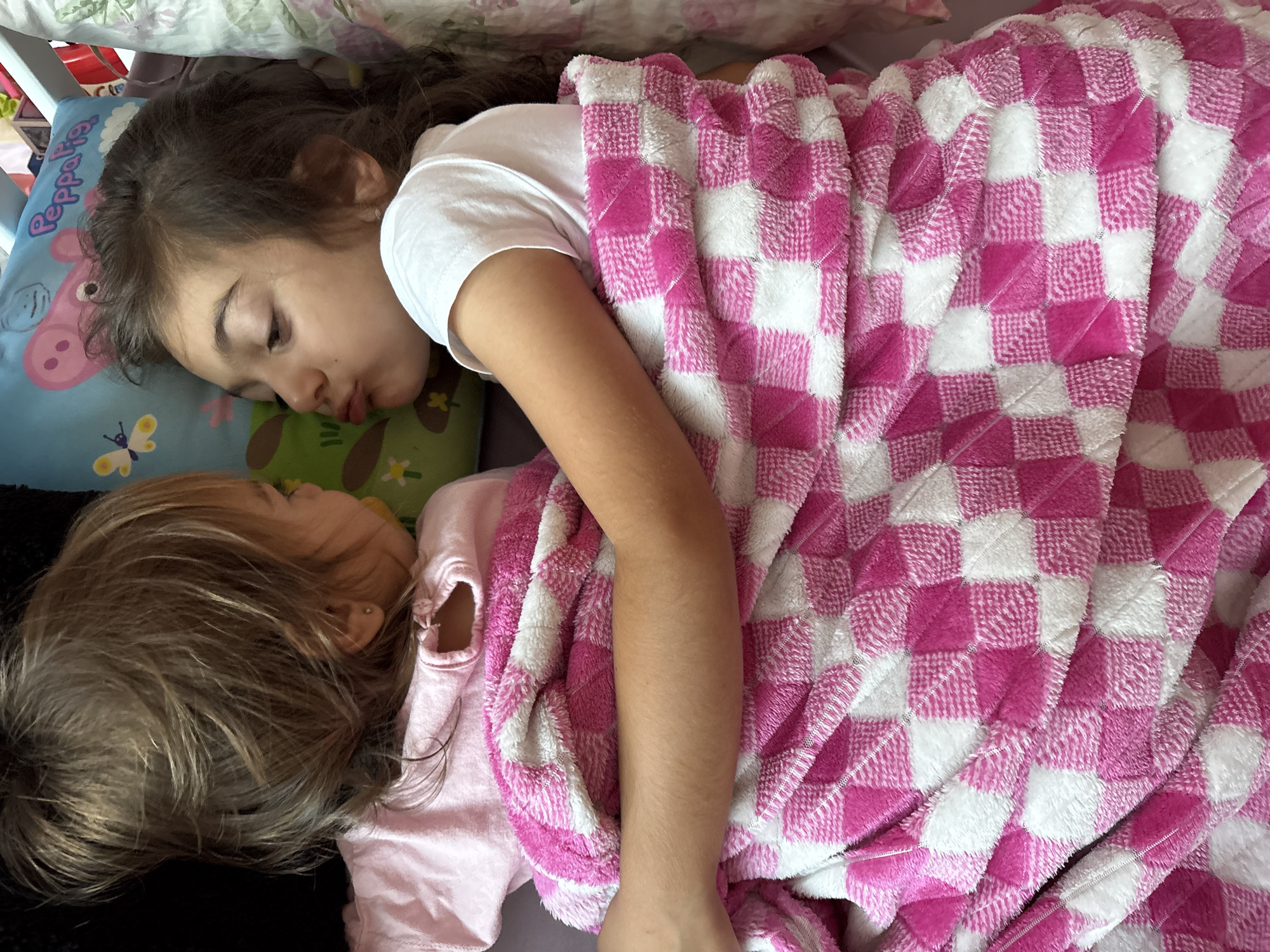 The image shows two young girls lying down, partially covered by a pink and white checkered blanket. The girl on the right has her lips puckered as if to kiss the girl on the left, who is looking away. Both girls are wearing t-shirts. The background includes a pillow with a cartoon character and text that appears to say "Peppa Pig". The scene suggests an indoor setting, possibly a bedroom or living area.