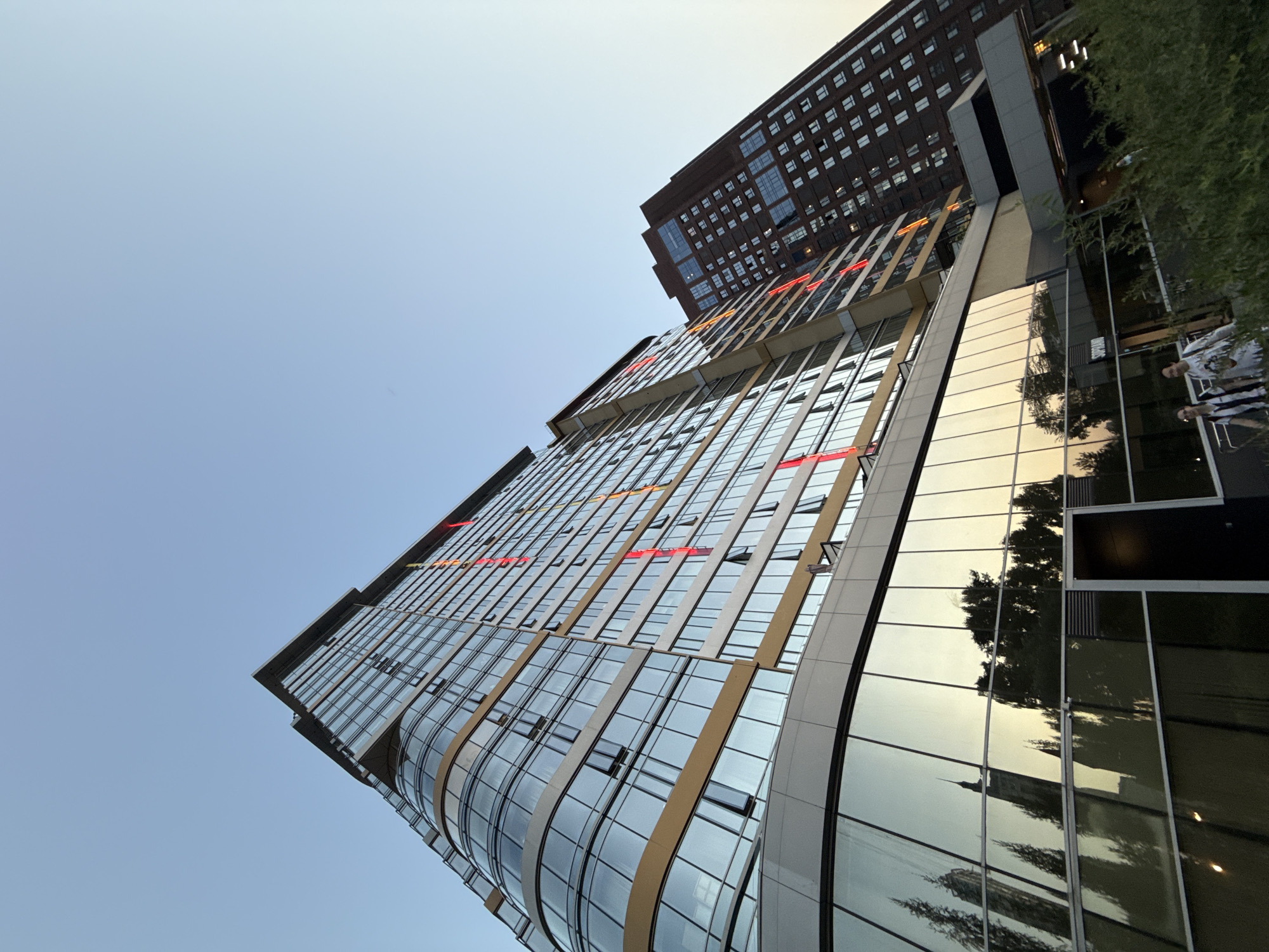 The image shows two modern high-rise buildings in Yekaterinburg, Russia. The building on the left is taller and has a curved glass facade with golden accents. Red and orange vertical lights are visible on the facade. The building on the right is made of dark brick with many windows. The reflection in the glass facade of the left building shows trees and other structures. In the foreground, two people are visible walking on a paved area. Lush green bushes are in the lower right corner of the image. The sky is clear and light blue, suggesting it is daytime.