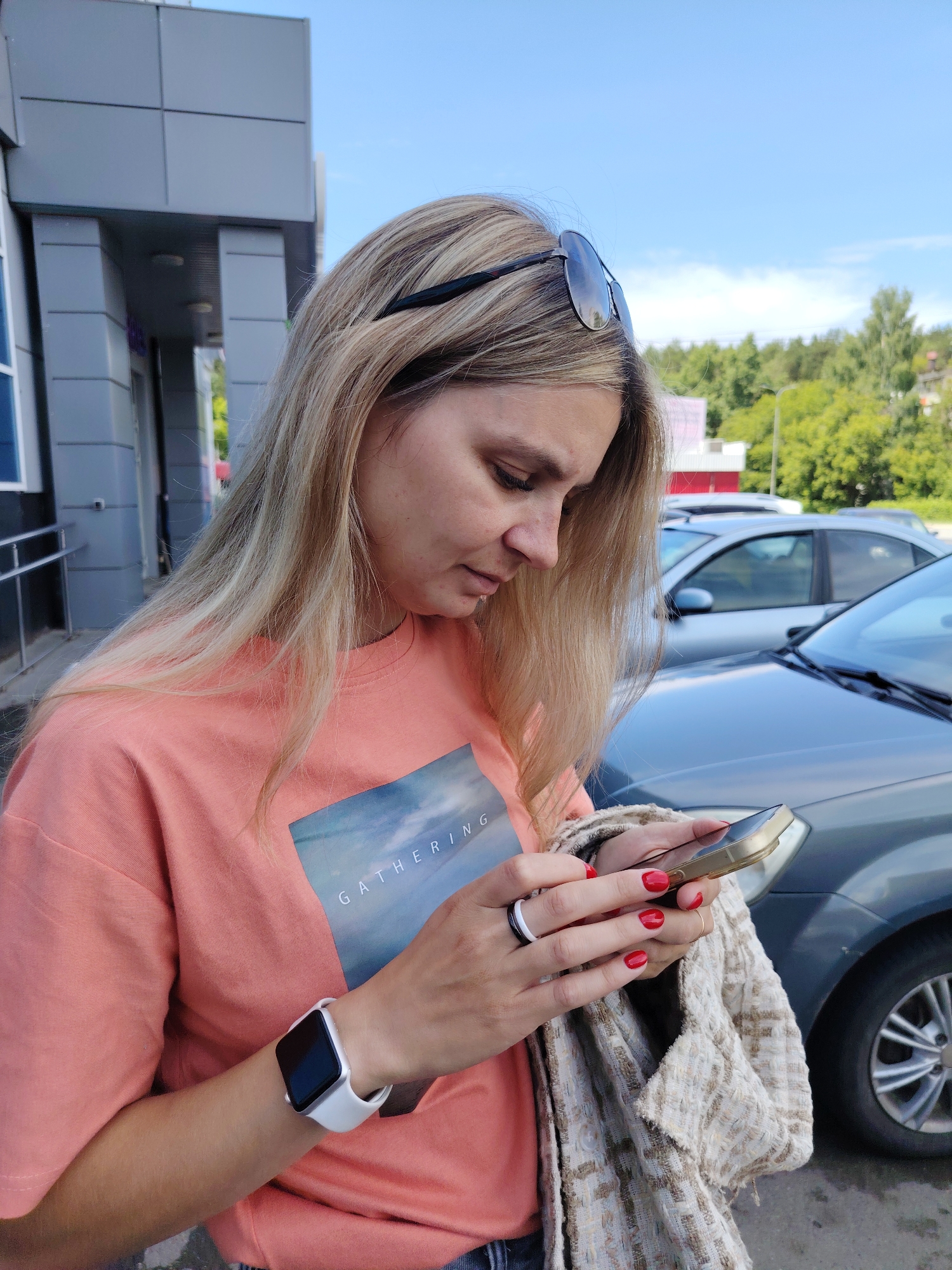 A blonde woman with sunglasses perched on her head looks down at her smartphone while holding a patterned jacket. She wears a coral t-shirt with the word "GATHERING" printed on it, and a smartwatch on her left wrist. Her fingernails are painted red. The woman is standing outdoors in what appears to be a parking lot, with cars and a building visible in the background. The sky is blue with a few clouds, and trees can be seen in the distance. The scene is likely located in Chaikovsky, Russia, based on contextual information not present in the image itself.