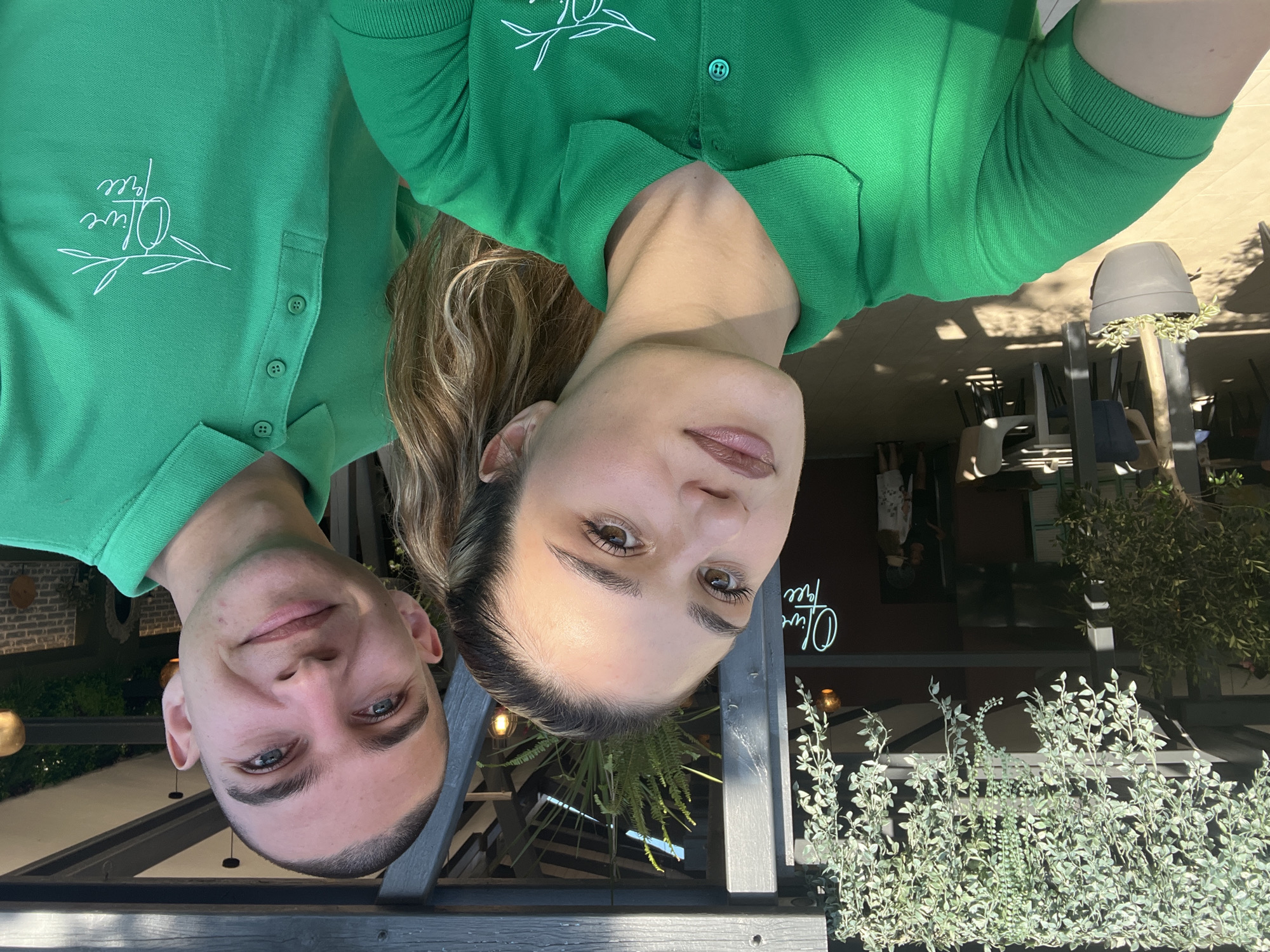 The image captures a selfie of a man and a woman, both wearing green polo shirts with the "Olive Tree" logo. The man, on the right, has a shaved head and blue eyes, smiling at the camera. The woman, on the left, has long blonde hair pulled back in a ponytail and is looking towards the camera with makeup on. They are standing outdoors, with lush greenery and a wooden structure visible in the background. The setting appears to be an outdoor seating area of a restaurant or cafe. In the background, there are tables and chairs, suggesting a dining environment. The lighting indicates it is daytime, possibly late afternoon due to the warm shadows. The presence of plants and the overall ambiance suggest a relaxed, possibly resort-like setting. The image does not contain any specific landmarks or identifying features that would place it definitively in Lozenets, Bulgaria.
