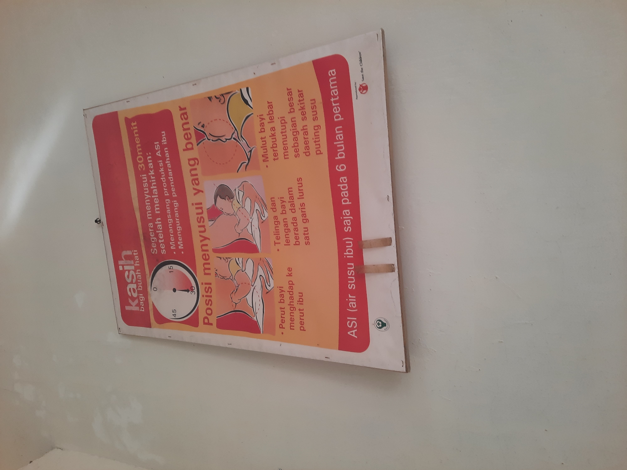 The image shows a poster on a white wall, possibly in a health clinic or educational facility in Lhokseumawe, Indonesia. The poster, titled "Kasih bagi buah hati" (Love for the little one), provides guidance on breastfeeding.
The poster features several key elements:
- A clock graphic indicating "0", "15", "30", and "45" minutes, suggesting a time frame related to breastfeeding.
- Text that reads "Segera menyusui 30menit setelah melahirkan:" (Breastfeed immediately 30 minutes after giving birth:) followed by bullet points:
- "Merangsang produksi ASI" (Stimulates breast milk production)
- "Mengurangi pendarahan ibu" (Reduces maternal bleeding)
- A section titled "Posisi menyusui yang benar" (Correct breastfeeding positions) with illustrations and accompanying text:
- An illustration of a baby being held close to the mother's chest.
- Text bullet points:
- "Perut bayi menghadap ke perut ibu" (Baby's stomach facing the mother's stomach)
- "Telinga, leher dan badan bayi berada dalam satu garis lurus" (Baby's ear, neck, and body in a straight line)
- "Mulut bayi terbuka lebar menutupi sebagian besar daerah sekitar puting susu" (Baby's mouth wide open covering most of the areola)
- The bottom of the poster has the text "ASI (air susu ibu) saja pada 6 bulan pertama" (Breast milk only for the first 6 months).
- A logo with a green cross and circle, likely representing a health organization, and the text "Save the Children" at the bottom right.
The wall appears to be slightly textured and shows some minor imperfections, with a shadow cast by an unseen light source from the top left. The poster itself is attached to the wall with what appear to be wooden planks on the bottom edge. The overall scene is clean and functional, typical of a public health information display.