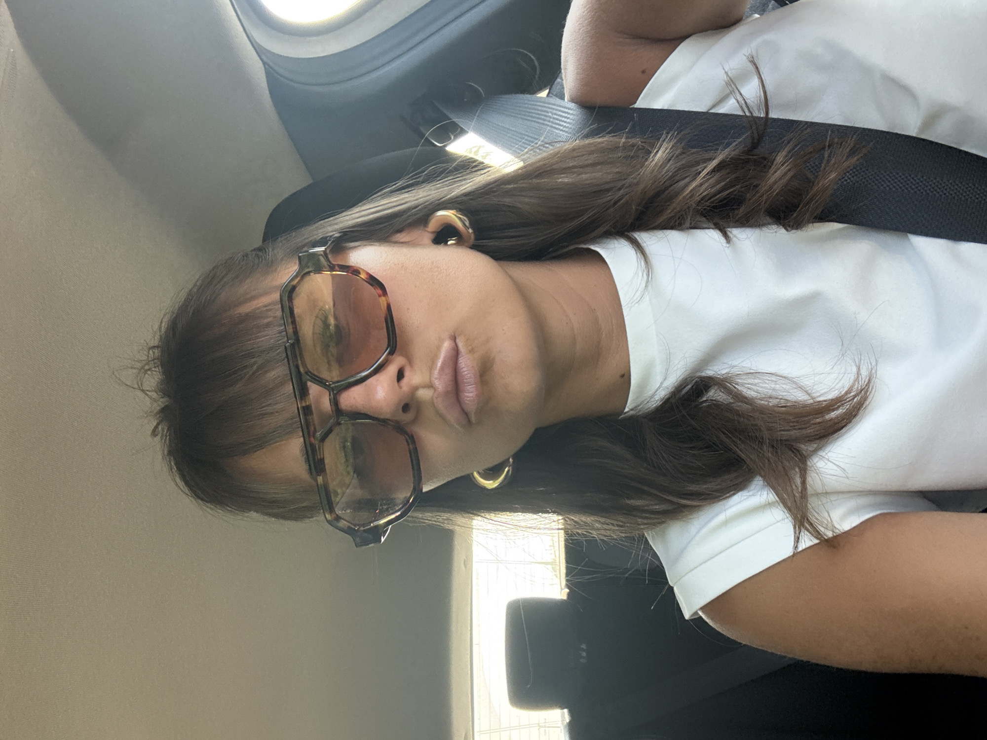 The image captures a woman taking a selfie or being photographed inside a car, set in Le Muy, France. She is seen from the chest up, positioned within the car's interior. She has long, straight brown hair styled with bangs, partially covering her forehead. She wears large, rectangular eyeglasses with a tortoiseshell-patterned frame and golden hoop earrings. Her lips are slightly pursed in a posed expression.
She is dressed in a plain white t-shirt, and a black car seatbelt is visible diagonally across her chest and shoulder. The background reveals elements of the car's interior, including a light-colored headliner or door panel on the left and a darker car seat or headrest on the right. Bright daylight streams through a car window in the upper background, indicating it is daytime. There is no visible text in the image.