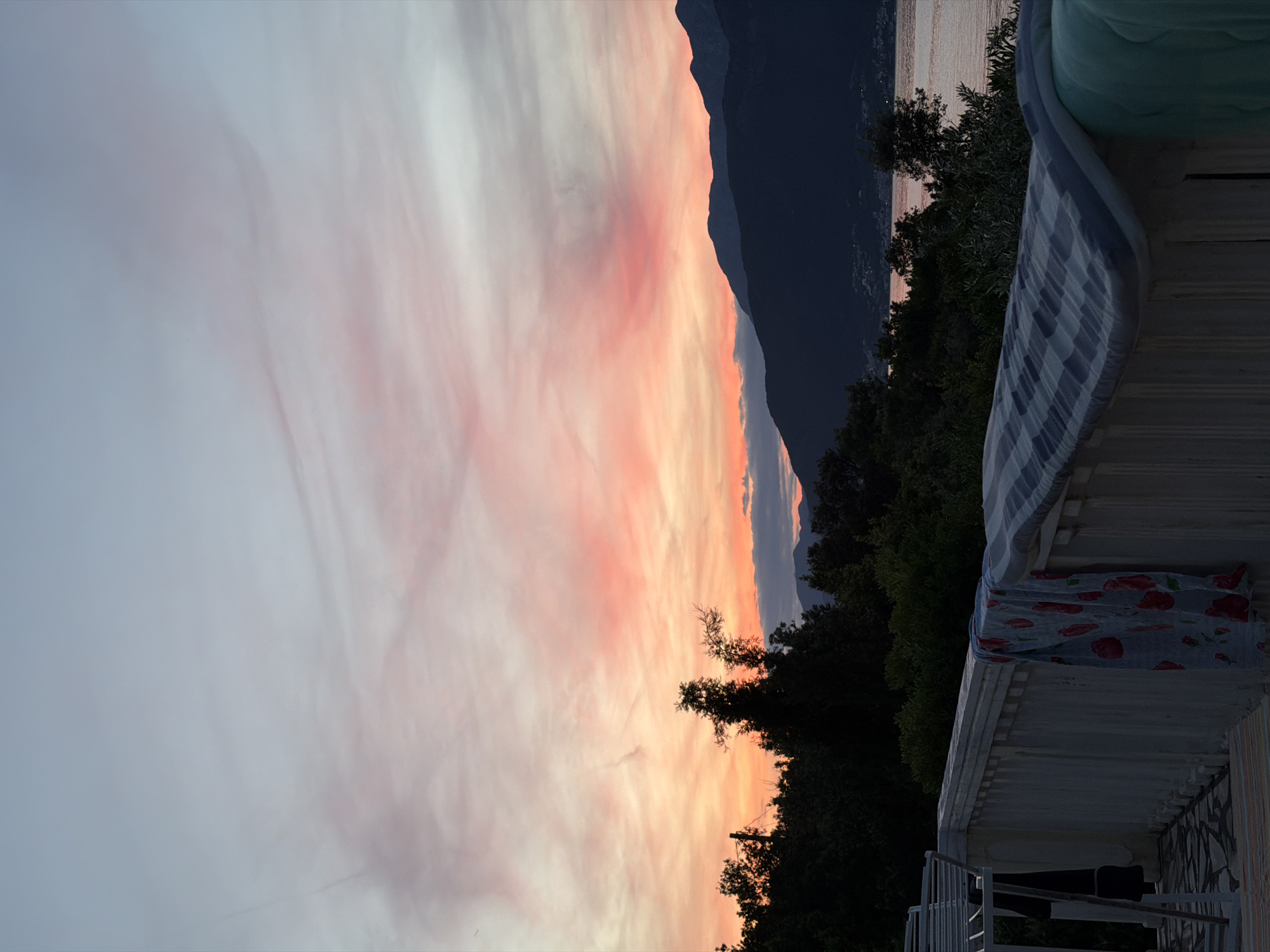 Here's a detailed description of the image:
The image captures a view of a sunset over the mountains and water, likely near the city of Gošići, Montenegro, based on the surrounding environment. The sky displays a gradient of colors from light pink and white near the top to deeper orange hues closer to the horizon. Fluffy clouds are scattered across the sky.
A mountain range dominates the middle ground, casting a dark, silhouetted shape against the vibrant sky. In the foreground, there is foliage, consisting of trees.
A structure with a white railing and blue and white awning is present in the lower-right corner of the image, partially obscuring the view.
