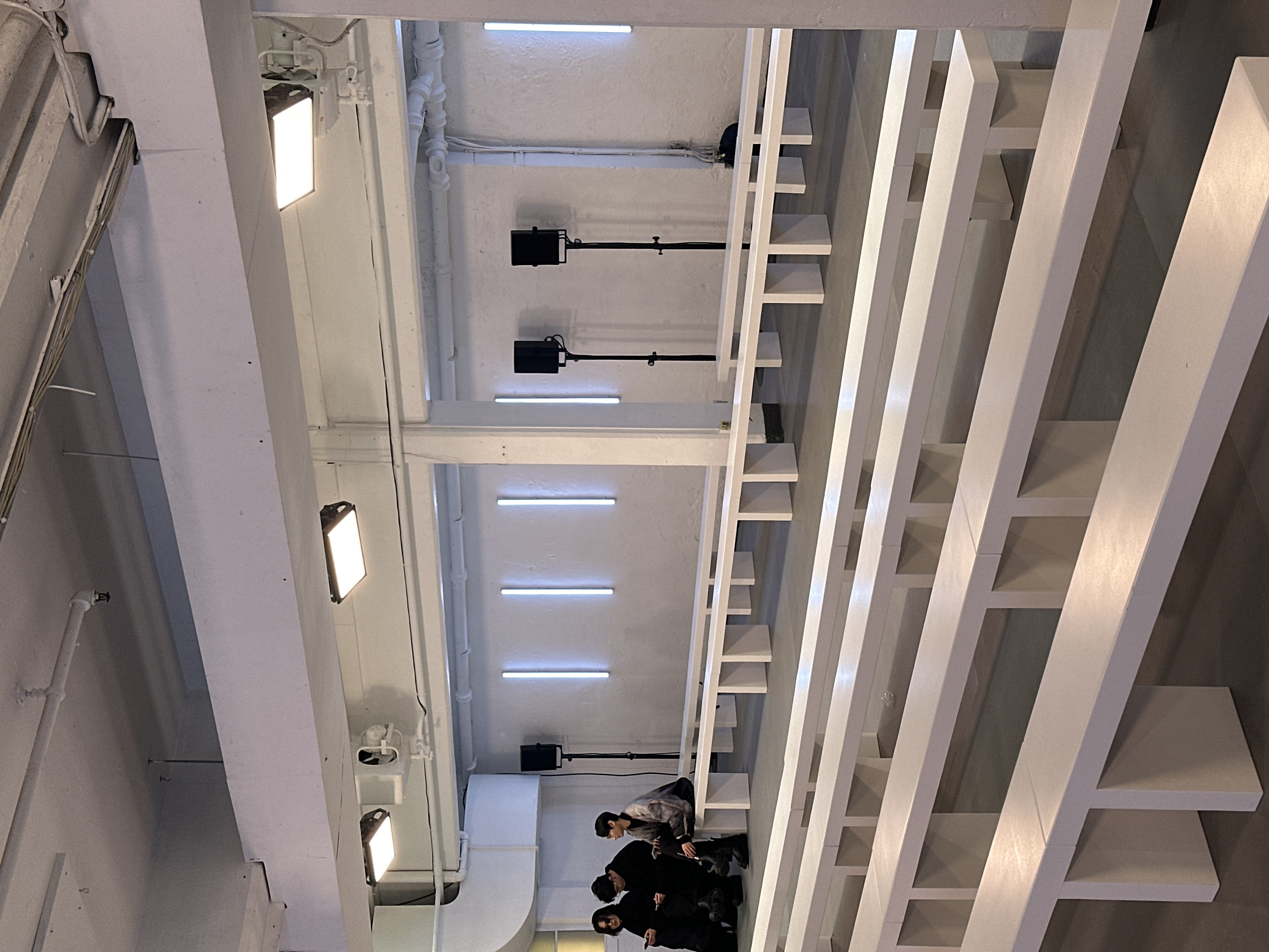 The image displays an interior architectural space characterized by a minimalist, industrial aesthetic, predominantly in white and light grey tones, illuminated by artificial light. The perspective is tilted, suggesting a dynamic upward or downward gaze within the space.
The environment features a ceiling and walls constructed from white or light grey concrete or plaster. Numerous white pipes, conduits, and a large ventilation duct are visible, running along the ceiling and walls, contributing to an exposed, industrial design. A fire sprinkler head is also noticeable on one of the pipes on the left.
The space is brightly lit by various artificial light sources:
* Several horizontal, rectangular LED strip lights are embedded or flush-mounted on the ceiling, casting a cool white light.
* Multiple rectangular, flat-panel LED lights are mounted on architectural elements, such as beams or columns on the left side, emitting intense white light.
* Two black, box-shaped spotlight-like fixtures are mounted on long, slender black arms extending from the ceiling in the mid-ground, suggesting directional or display lighting.
A prominent architectural feature dominates the right side of the frame: a large, geometric structure made of interlocking white beams and horizontal shelf-like projections. This structure creates a striking, abstract pattern and extends from the foreground into the background.
In the lower right section of the image, beneath this white geometric structure, three individuals, likely young adults, are visible. They appear to be observing something outside the frame or resting.
* One person, wearing a light-colored, patterned long-sleeved shirt and dark pants, is seated or leaning on one of the white shelf-like projections of the geometric structure.
* The other two individuals are standing below the first person, dressed in dark (mostly black) clothing, including dark shirts and pants/boots. One of them appears to be holding a small, dark object. All three are looking in the same general direction.
The overall scene suggests a contemporary art gallery, a showroom, a modern retail space, or an event venue. The clean lines, bright lighting, and monochromatic palette create a modern and somewhat stark atmosphere. There is no visible text in the image.