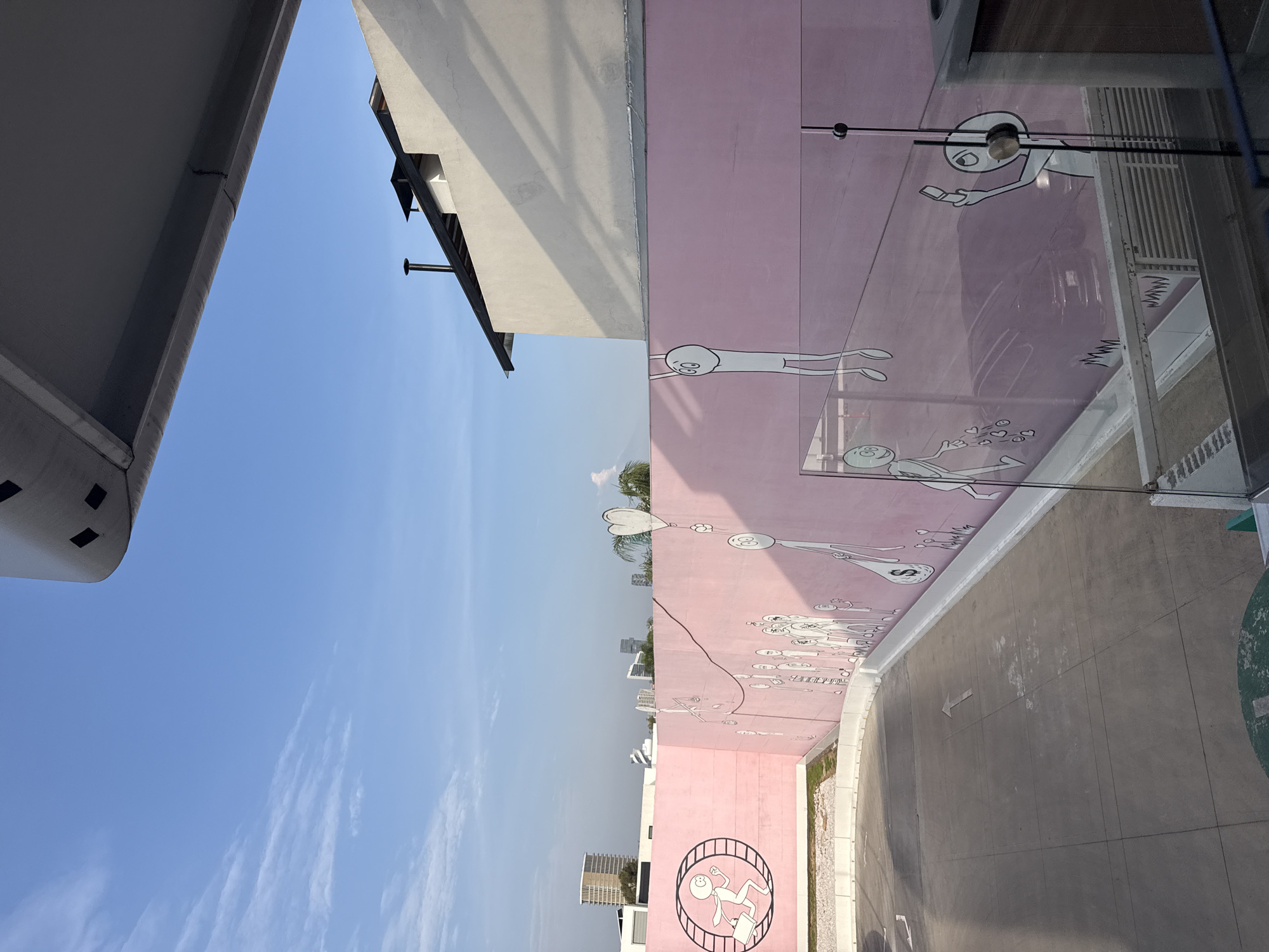 The image depicts an outdoor urban scene in daylight, likely in Puebla, Mexico. A large, light pink building with an extensive mural dominates the right side, while a simpler, light-colored building structure is visible on the left, under a clear blue sky with sparse white clouds.
The mural on the pink building features numerous whimsical, stick-figure-like characters in black outline. At the top right, a character appears to be holding a smartphone. Below, another figure is surrounded by hearts and other small shapes, while further down, a character holds a heart-shaped balloon attached by a string. A long line of standing figures extends horizontally across the middle of the wall. Towards the bottom, a figure is depicted running inside a hamster wheel, clutching a briefcase. Other figures are shown in various dynamic poses, some seemingly floating or falling, connected by thin lines. A money bag symbol with a dollar sign is visible near some characters. Glass panels or railings are present along the top edge of the pink wall, reflecting parts of the mural.
The foreground consists of a paved, grey concrete area, possibly a street or parking lot, extending from the base of the pink building. A faded white arrow is painted on the ground, pointing away from the wall. A circular patch of green ground is also visible in the bottom right corner. The background reveals additional urban buildings in the distance, indicating a city environment. Sunlight casts shadows on the buildings and ground, suggesting a bright and sunny day. No explicit text is clearly visible other than the ground arrow.