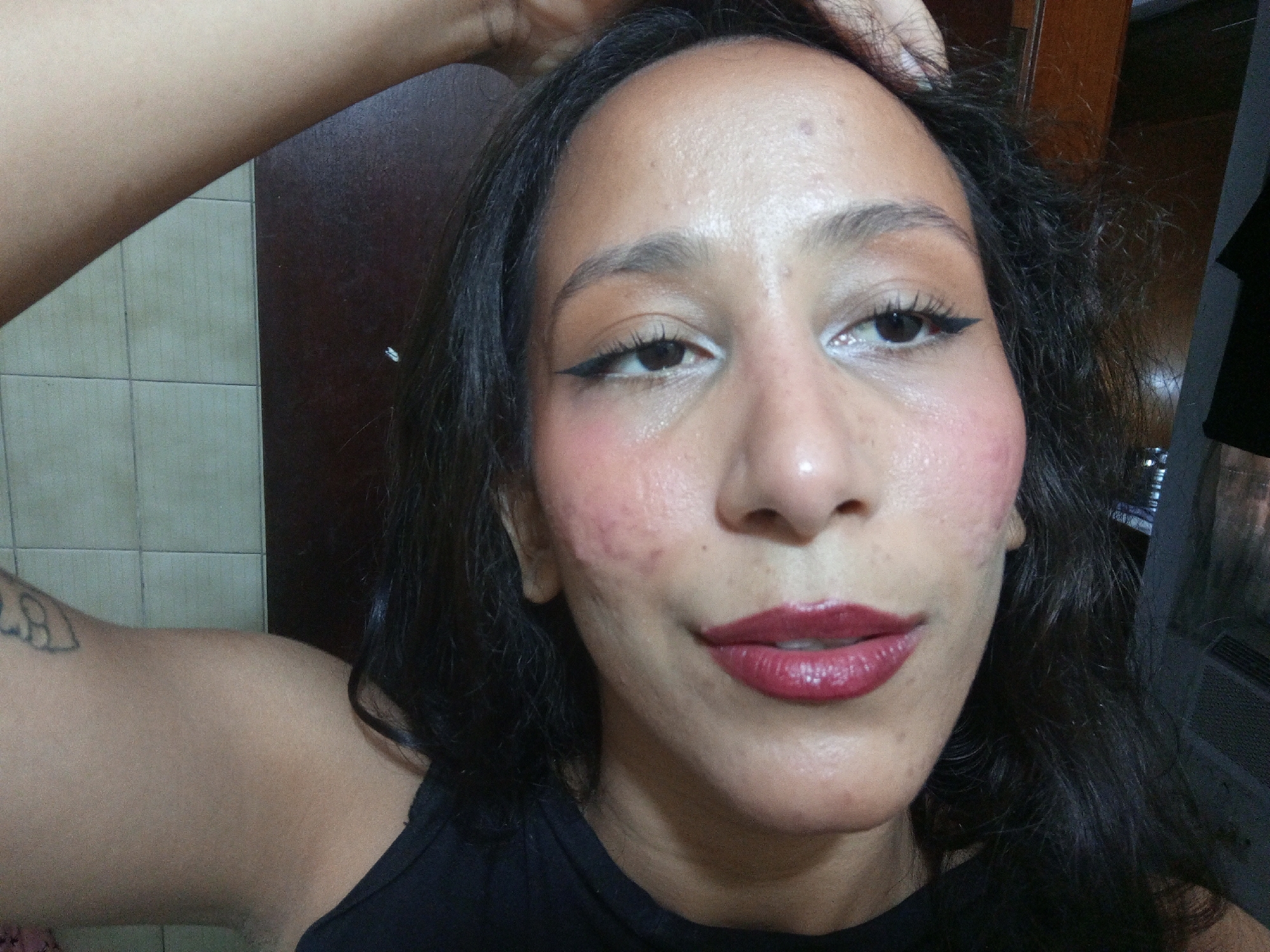 A woman is taking a selfie indoors. She has medium brown skin, dark hair, and is wearing makeup, including eyeliner and dark red lipstick. She is wearing a black top. Her right arm is raised with her hand touching her hair. Part of a tattoo is visible on her upper left arm. Behind her is a dark brown door and tiled wall. Part of a window air conditioner is visible in the background on the right. The city of Porlamar, Venezuela cannot be determined from the image.
