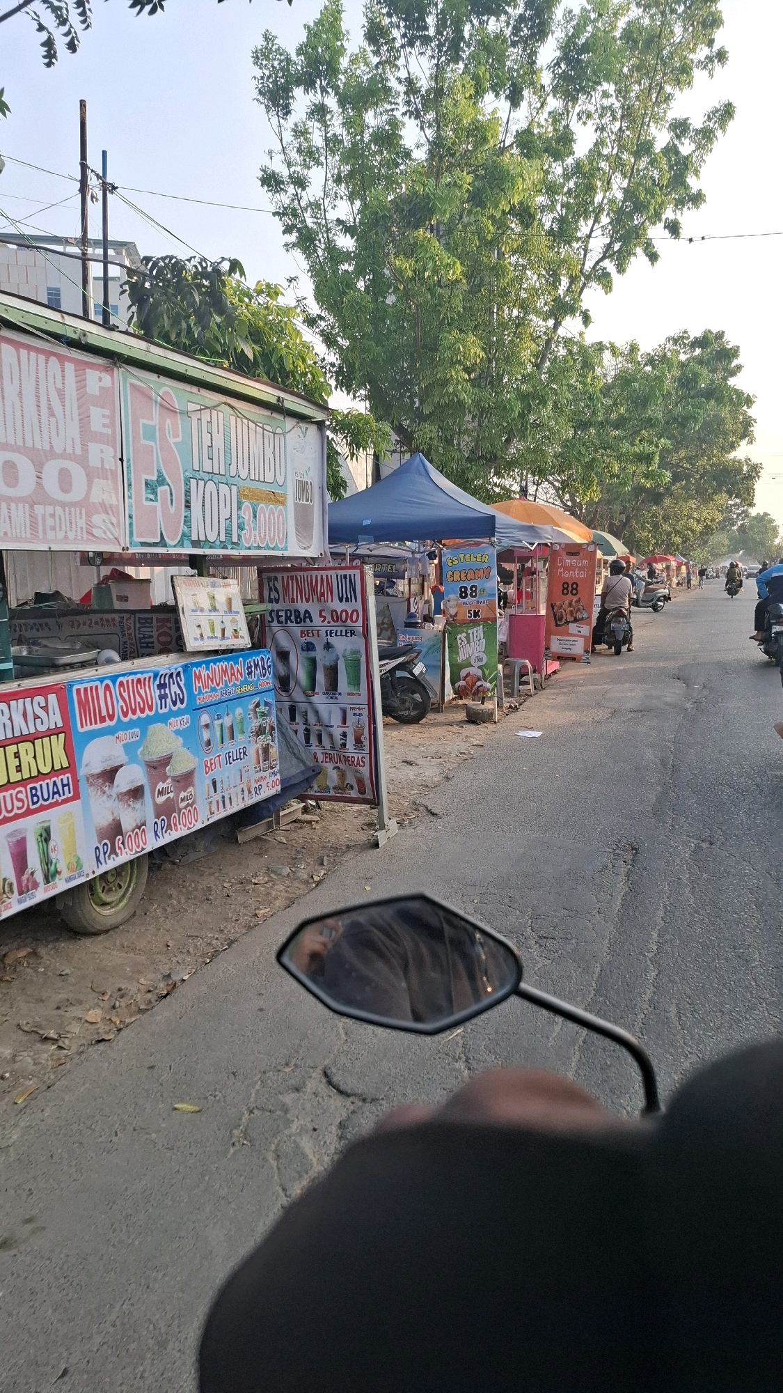 An outdoor, ground-level, eye-level medium shot shows a street-side food stall with a variety of drinks and snacks being sold. The stall is set up along a paved road in Bandar Lampung, Indonesia. The focus is on a food cart displaying colorful signage advertising various drinks, including "MILO SUSU," "ES TEH JUMBO KOPI," and "ES TEH KERAMY." Several drinks are pictured on the signs, some in clear cups with straws. Next to the food cart, a blue tent provides shade. Further down the road, other stalls are visible, some with umbrellas and awnings. Several people are on motorcycles, either parked or driving past. One person is seen riding a motorcycle in the distance, and another is partially visible in the foreground, reflected in a motorcycle's rearview mirror. The scene is illuminated by natural sunlight, creating a bright atmosphere. Trees with green foliage line the background, and power lines are strung overhead. The road itself appears to be in fair condition, with some visible cracks and patches. The overall impression is that of a bustling local market or roadside eatery.