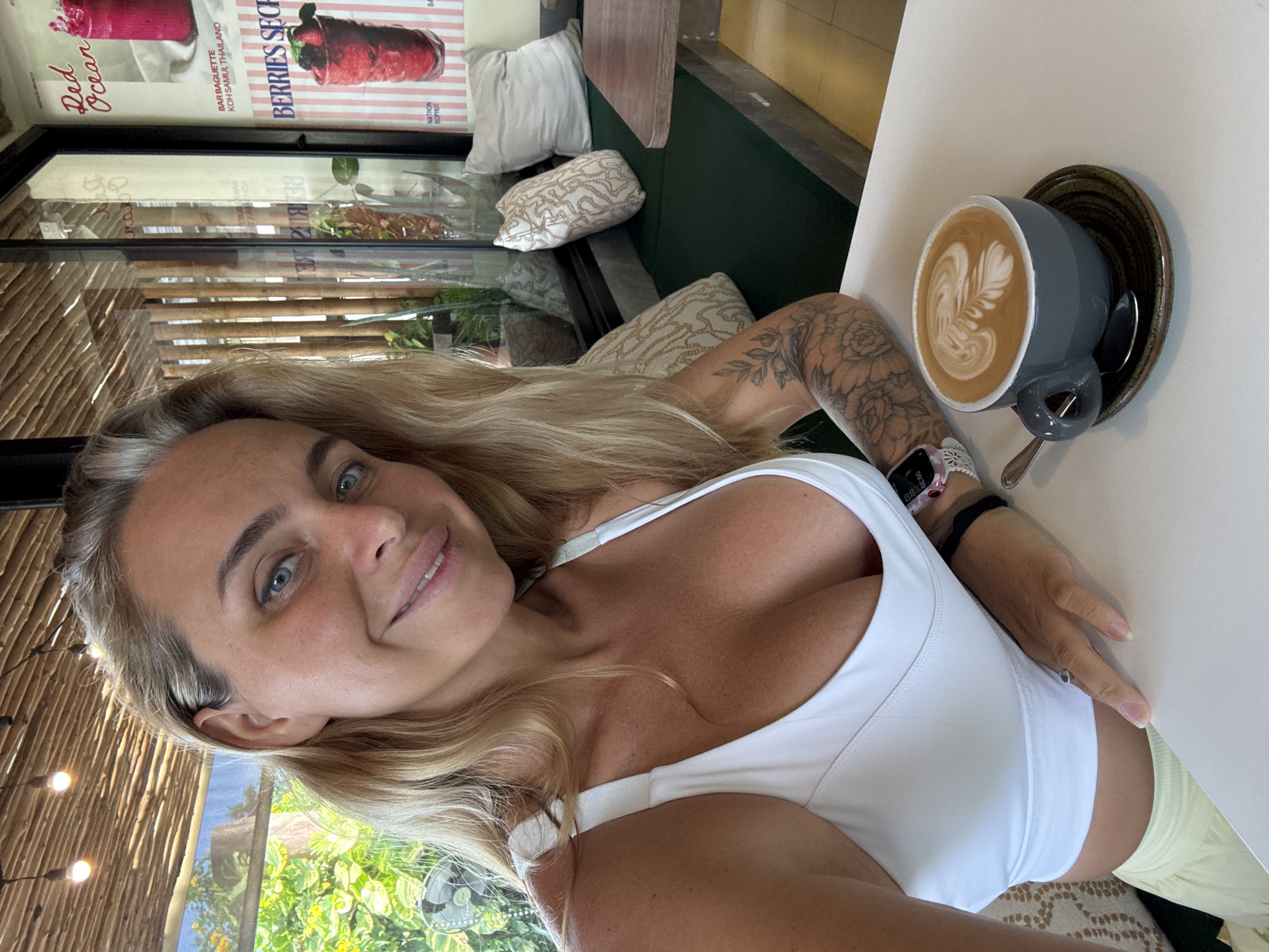 A woman with blonde hair and blue eyes sits at a table in a cafe in Koh Samui, Thailand. She is wearing a white sports bra and yellow pants. A cup of coffee with latte art is placed on the table in front of her. The cafe has a rustic interior with bamboo accents and large windows that overlook lush greenery. Posters advertising drinks are visible on the wall.