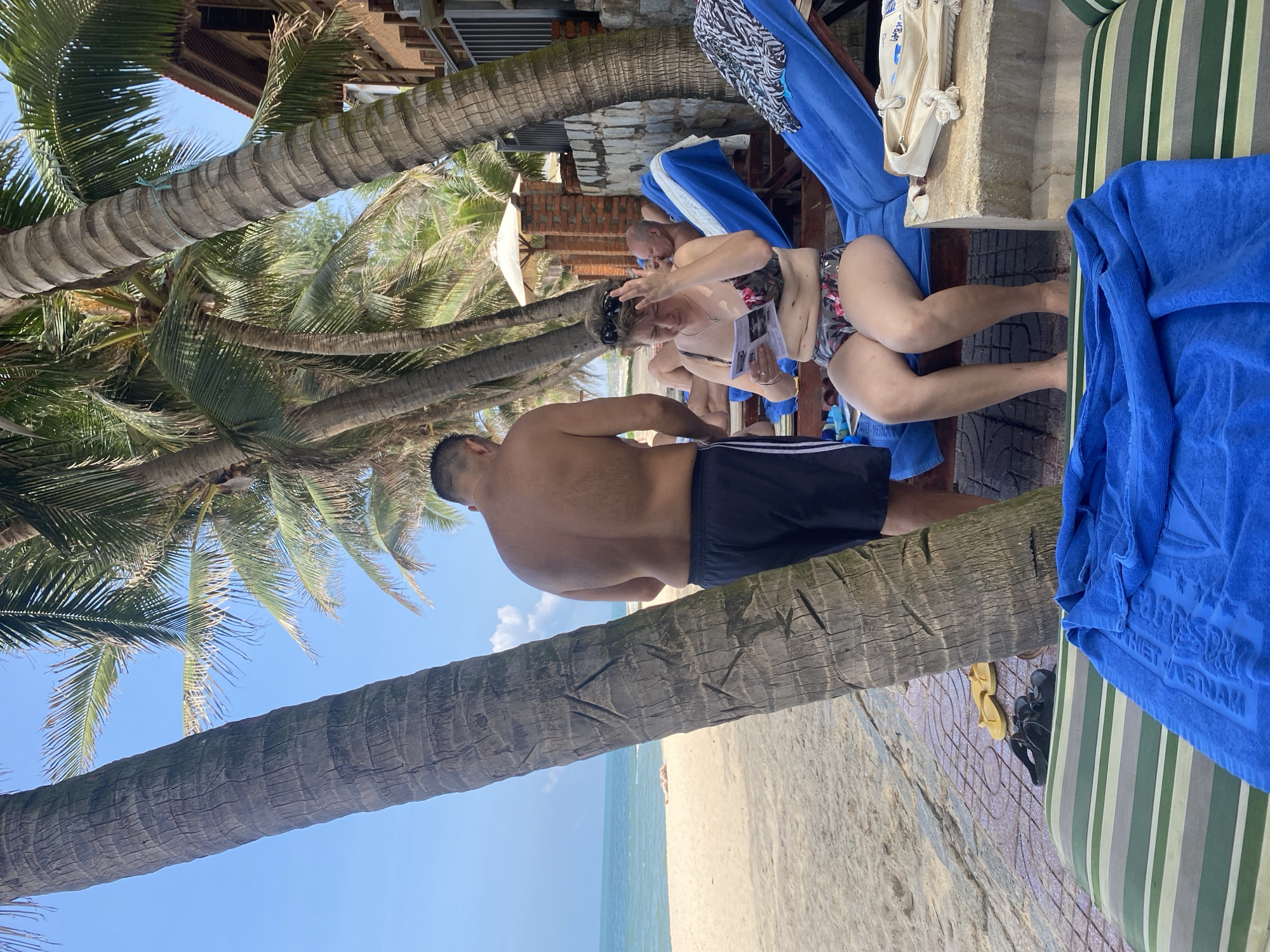 This image captures an outdoor scene at a beach resort in Phu Thuy Ward, Vietnam. The environment is dominated by a clear blue sky with sparse white clouds, a sandy beach, and a calm blue-green ocean to the left.
In the foreground, a large, rough-textured palm tree trunk occupies the left side, displaying several carved markings. At its base, on a patterned paved surface, a pair of yellow flip-flops and a pair of dark sandals are placed. A partially visible lounge chair with a green and white striped cushion is positioned in the immediate foreground, draped with a folded blue towel that appears to have "VIET NAM" embroidered on it.
To the right of the prominent palm tree, a man stands with his back mostly towards the viewer. He wears dark swim shorts, and his upper torso is bare. To his right, a woman sits on a blue lounge chair, facing slightly towards the left. She wears a floral two-piece swimsuit, and dark sunglasses are pushed up onto her head. She is engaged in reading an open white booklet or brochure. Adjacent to her lounge chair, on a light-colored stone surface, a canvas beach bag with rope handles is situated. On another blue lounge chair positioned further to the right, a black and white patterned towel is draped. Behind the sitting woman, a second man is visible, lying on a blue lounge chair. He has a bald or very closely shaven head, and his right hand is raised to his forehead.
The midground and background feature multiple tall palm trees with green fronds, providing shade. One palm tree trunk has a blue rope tied around it. Further back, a white beach umbrella is visible between some palm trees and low brick walls. Beyond these, a multi-story building with a reddish-brown roof and wooden architectural details is partially seen on the far right, suggesting a hotel or resort structure. The area beneath the lounge chairs is a paved surface, which transitions to light-colored sand leading to the ocean. In the distant background on the sandy beach, a single, prone figure is discernible near the waterline. The scene is brightly lit by natural sunlight, casting shadows.