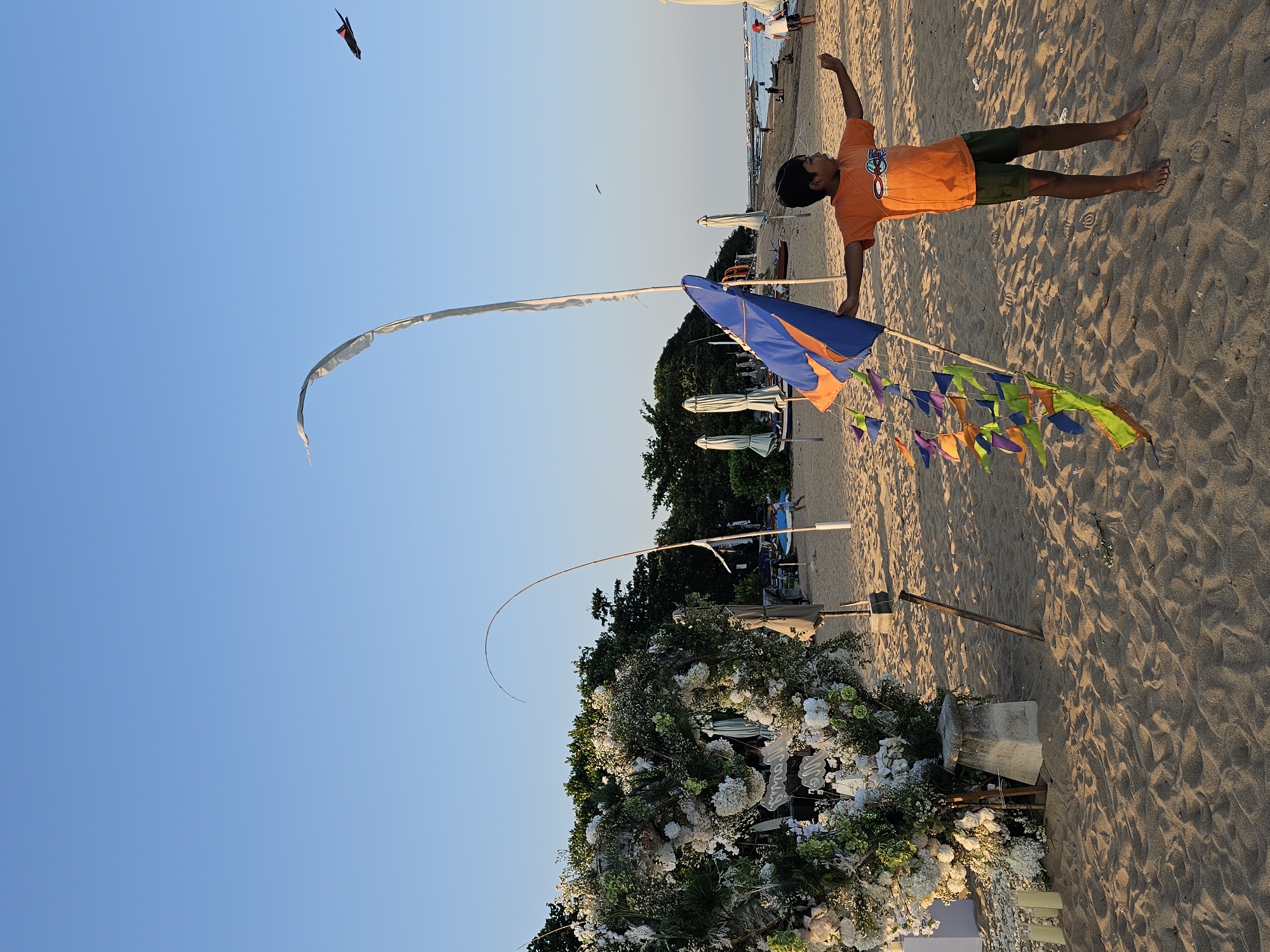 The image captures a beach scene near Denpasar, Indonesia, under a clear blue sky. A young boy wearing an orange t-shirt and green shorts is lying on his side on the sand, appearing to be in motion, possibly flying a kite. The foreground features a floral archway decorated with white and green flowers, suggesting a beach wedding or event. A tall, thin bamboo pole is erected behind the arch, adorned with a blue and orange flag, as well as colorful pennants. Another curved bamboo pole extends towards the sky. In the background, other beachgoers, umbrellas, and boats are visible along the shoreline. In the distant sky, a black kite is soaring.