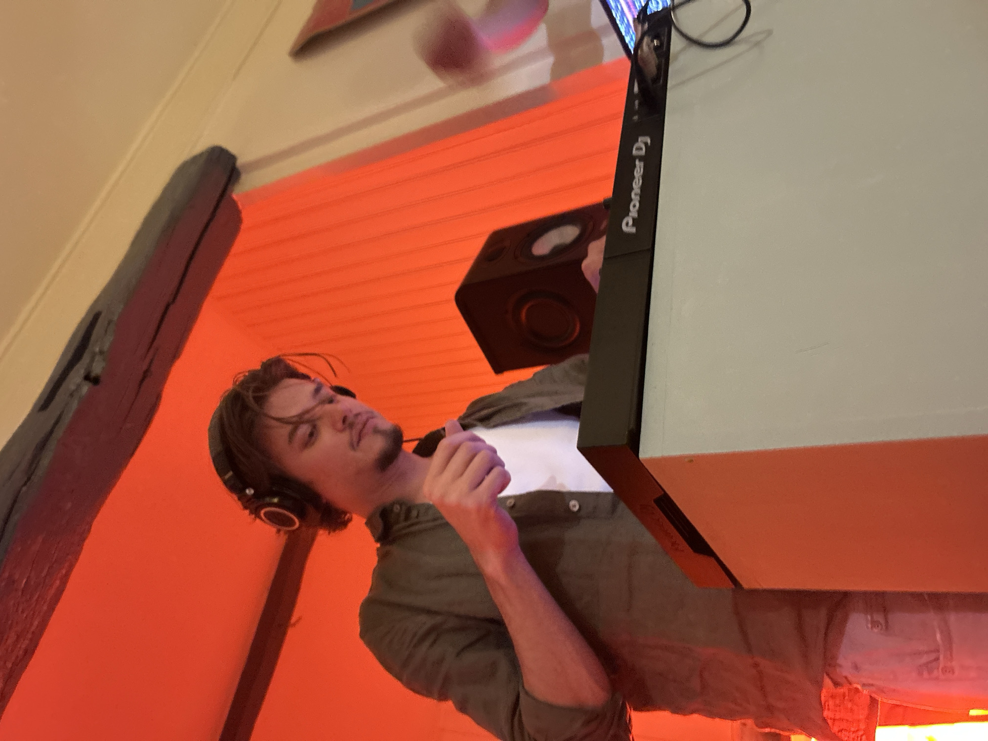 A person with brown hair and headphones is standing near a Pioneer DJ sound mixing console and a black speaker. The room has orange-toned lighting. A dark wooden beam is visible near the left edge of the frame. The city and specific location are unknown.
