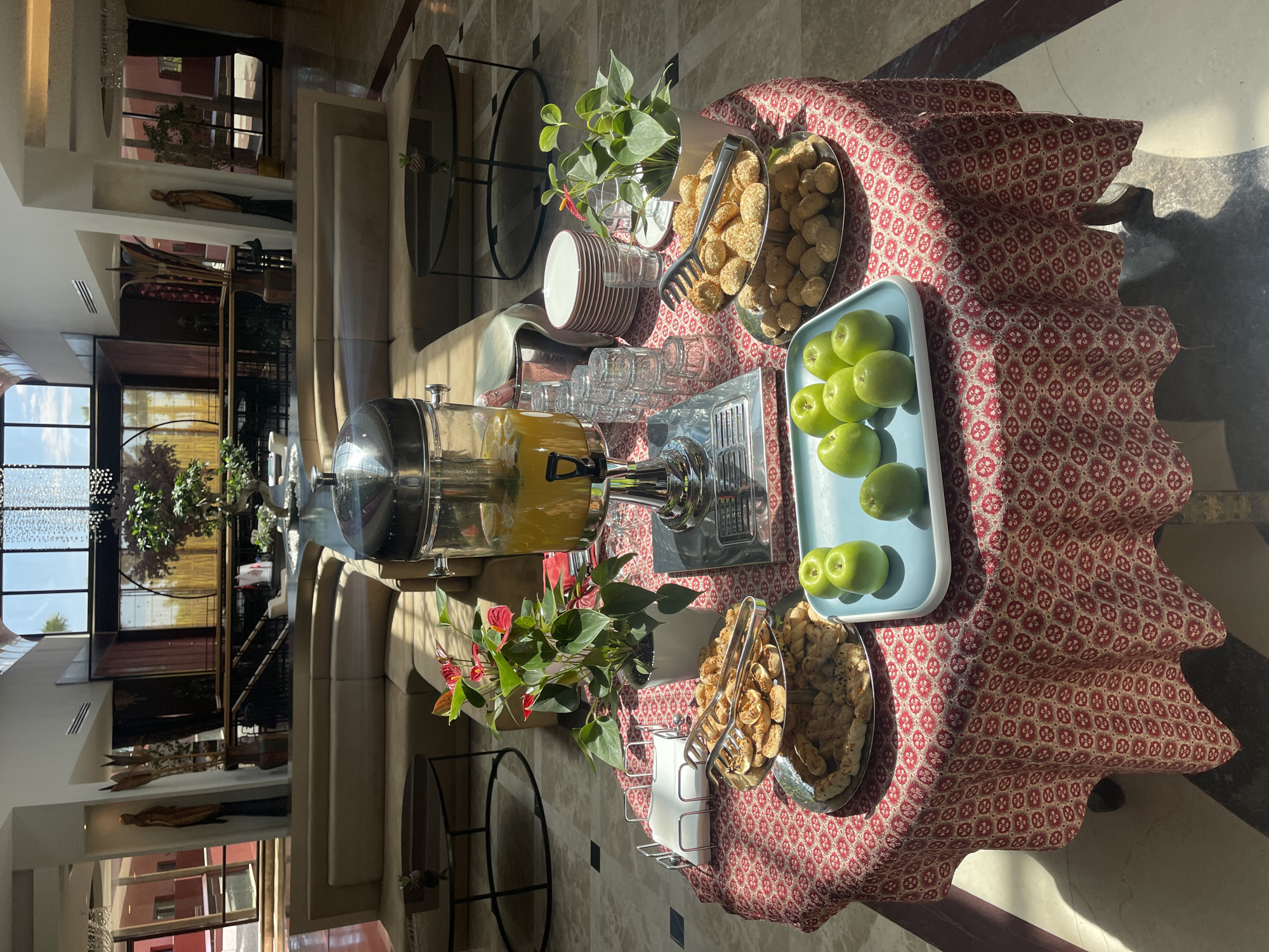 The image depicts a table set with refreshments in what appears to be a hotel lobby or lounge area, possibly in Unknown, Unknown. On the table, which is covered with a red patterned tablecloth, there is a drink dispenser filled with orange juice, surrounded by glasses, and trays of apples and cookies. Plants in white vases add a touch of decor. The setting is well-lit, with natural light coming through windows in the background, which overlooks a multi-level seating area. The architecture suggests a modern or contemporary design, with clean lines and open spaces. The scene seems quiet and inviting.