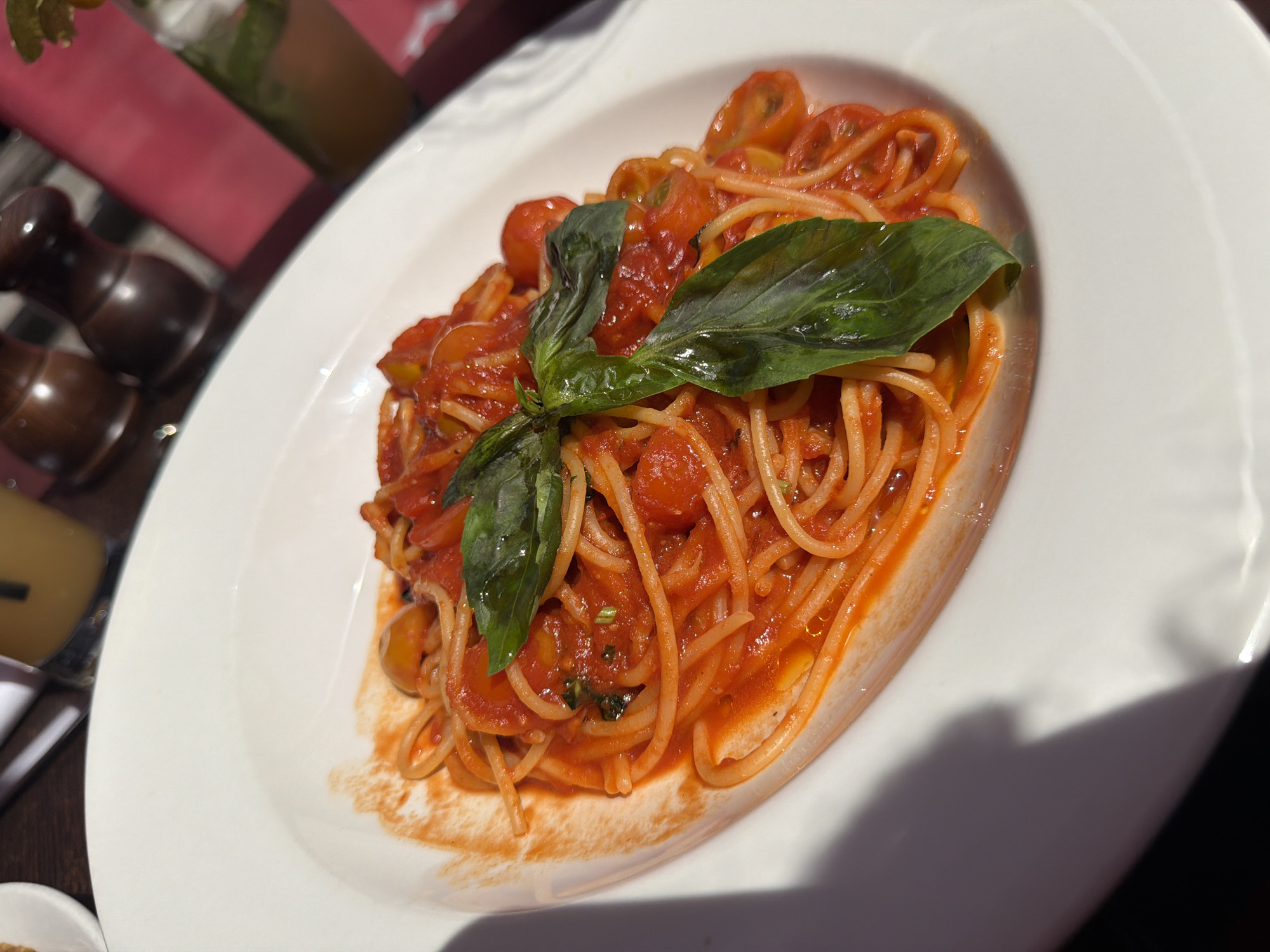 A plate of spaghetti with tomato sauce and basil sits on a white plate. The dish appears to be served outdoors, potentially in a restaurant, given the presence of a salt and pepper grinder and a drink in the background. The location is Guildford, United Kingdom. The plate is positioned at an angle. The sauce includes halved cherry tomatoes. Three basil leaves garnish the dish.