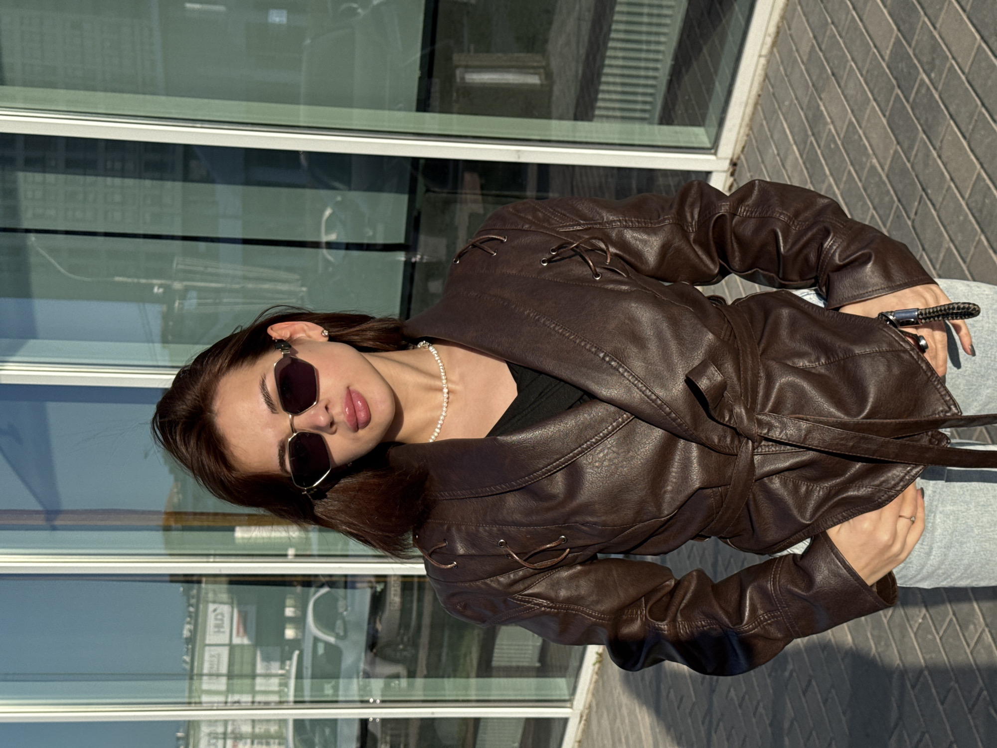 The image depicts a young woman outdoors, wearing a brown leather jacket, sunglasses, and a pearl necklace. She is standing in front of a modern building with glass walls that reflect the surrounding urban environment. The building appears to be located in Perm, Russia, based on reflections in the glass that show recognizable Cyrillic script signage. The woman is holding something in her hands, possibly a phone or keys. She is positioned on a paved area, likely a sidewalk or plaza, adjacent to the building. The time of day is daytime, indicated by the natural lighting and shadows. The photograph is taken at an angle, giving a tilted perspective of the scene.