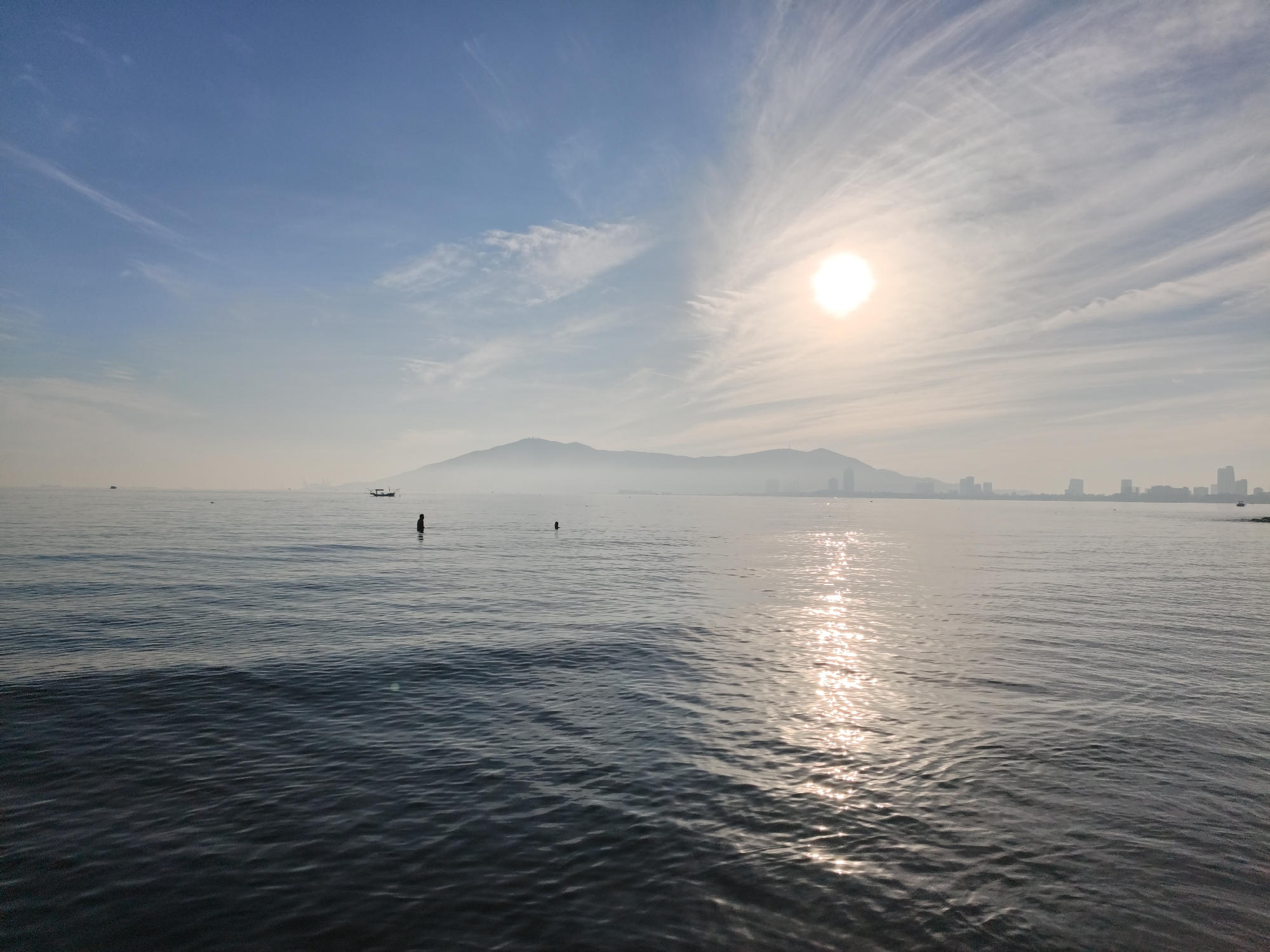 The image depicts a coastal view of Đà Nẵng, Vietnam, likely taken from a beach or a waterfront area. In the foreground, the calm sea displays gentle ripples, reflecting the bright sunlight. The sun, a luminous orb, is positioned high in the sky, partially obscured by wispy, feathery clouds that spread across the blue expanse. In the middle ground, several small boats are visible on the water. One of these boats appears to be a traditional fishing vessel. Two individuals are standing in the water, one appearing to be deeper in the sea than the other. In the distance, a hazy mountain range forms a silhouette against the horizon, with a city skyline peeking through the mist at its base. The city's buildings suggest a densely populated urban area adjacent to the natural landscape. The overall atmosphere is serene and tranquil, characteristic of a bright day by the sea.