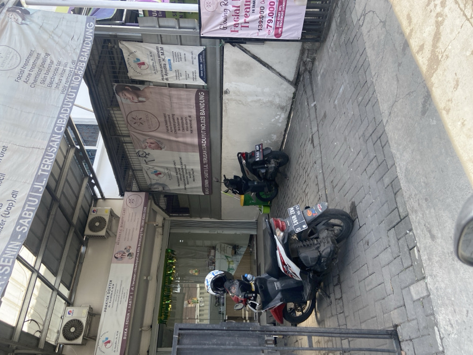 The image shows a storefront in Kecamatan Dayeuhkolot, Indonesia, located at latitude: -6.967, longitude: 107.592. The storefront has a sign that reads "Praktek Dokter dr. Ayudita, H, M,M" and "Aesthetic Beauty Care." Two motorcycles are parked in front of the storefront. There are multiple signs advertising beauty treatments and services, with prices listed. The storefront appears to be a beauty clinic or spa. The environment is an urban area, with paved sidewalks. The motorcycles are parked in the shade of a canopy, and the storefront has a metal gate. The signs are in Indonesian and English. The image is taken from a slightly elevated perspective.