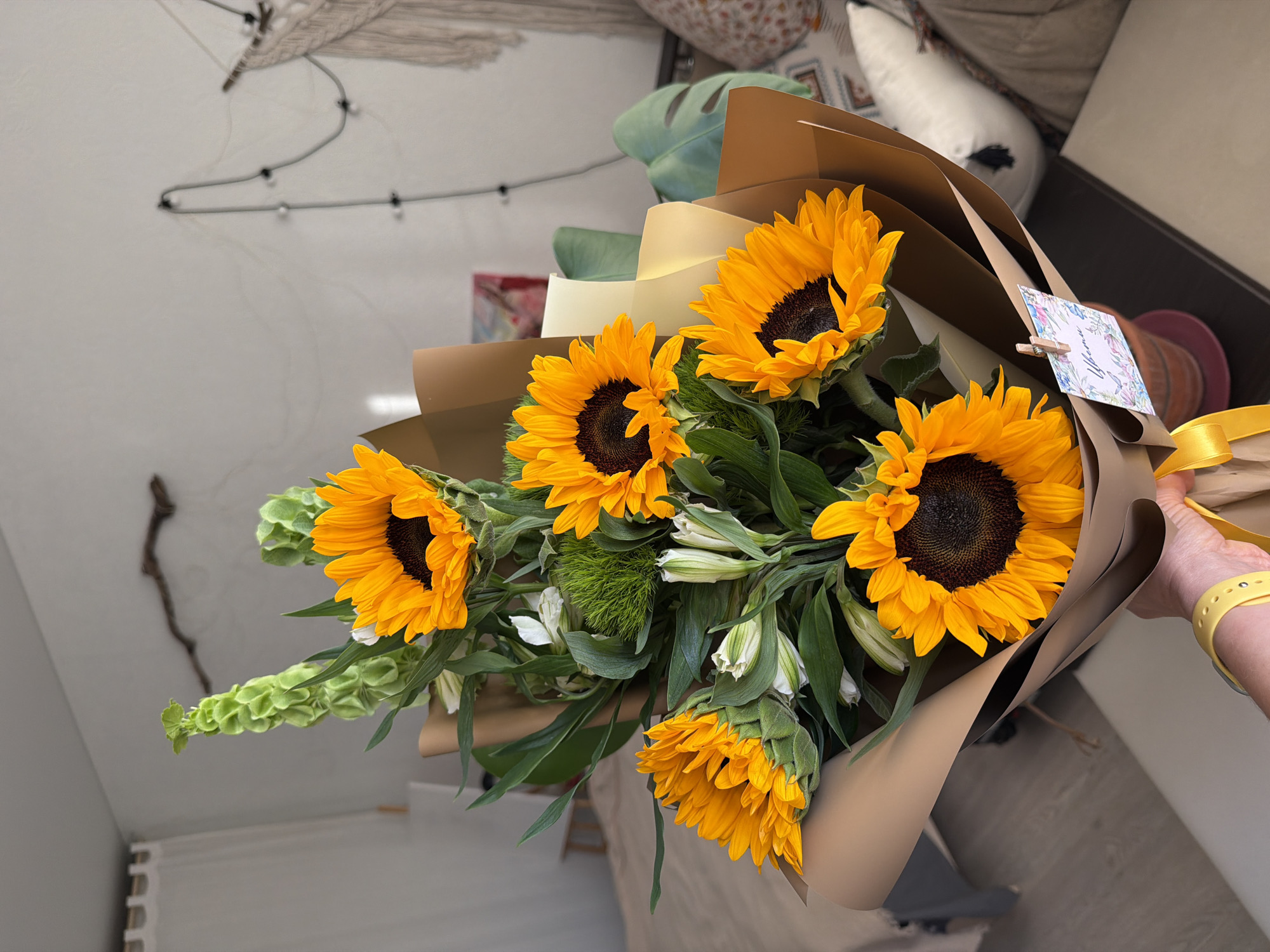 A bouquet of sunflowers and green flowers wrapped in brown paper is being held by a person with a yellow watch. The bouquet contains at least five large sunflowers with dark brown centers and bright yellow petals. There are green, leafy filler flowers and some white flowers interspersed among the sunflowers. The bouquet is wrapped in several layers of brown paper, and there is a yellow ribbon tied around the base. A small card is attached to the bouquet with a miniature clothespin. The person holding the bouquet has pale skin and a yellow watch on their wrist. The background is a blurry room with a macrame wall hanging and patterned pillows.