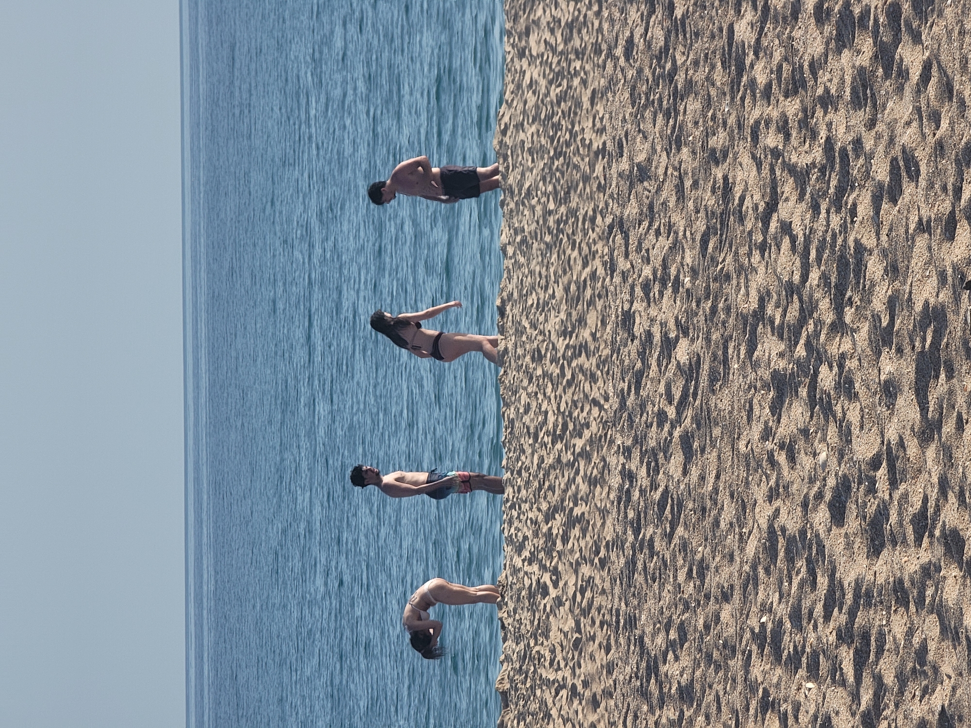 Four individuals are positioned on the sand near the ocean's edge. From left to right, the first person, a female, is bent over with her head down. The second person, a male, stands facing slightly to his right. The third person, a female, stands facing to her left. The fourth person, a male, stands facing to his left. The scene takes place on a sandy beach with the blue ocean in the background. The sky above is clear and light blue. The location is Quarteira, Portugal.