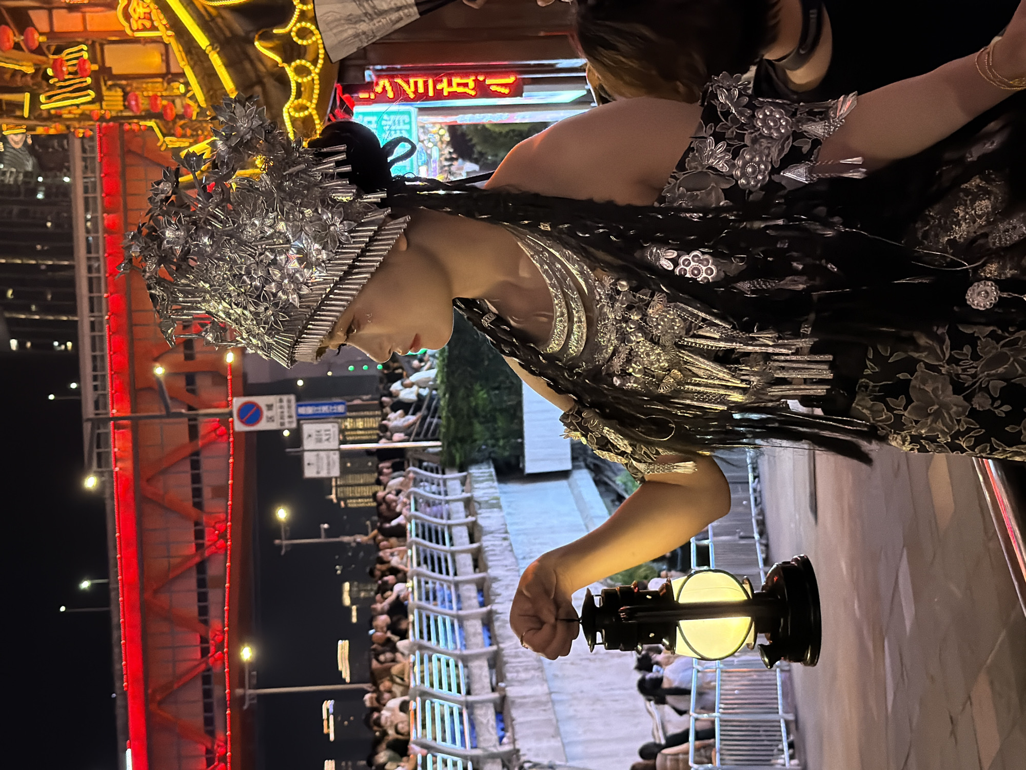 A person adorned in an elaborate silver headdress and a dark, intricately decorated costume is the primary subject of this image. The costume features floral motifs and metallic embellishments. The individual is shown from the chest up, with their head turned to the side, looking downwards. They are holding a vintage-style lantern in their right hand, which is illuminated. The scene appears to be at night, indicated by the artificial lighting and the dark sky.
The background suggests an outdoor urban setting, possibly a street or a promenade. A bright red illuminated bridge or structure is visible to the left, along with buildings and other lights in the distance. There is a crowd of people in the background, suggesting an event or a busy public space. A railing with a decorative pattern runs along the right side of the frame, and further back, there are more people and structures.
The overall mood seems festive or ceremonial, given the elaborate attire and the gathering of people. The lighting, with the contrast between the bright red bridge and the ambient streetlights, creates a vibrant atmosphere. There are some street signs visible, but the text is not clearly legible. The presence of the lantern and the ceremonial costume hints at a possible cultural or religious event. The location context suggests Yuzhong District, China.