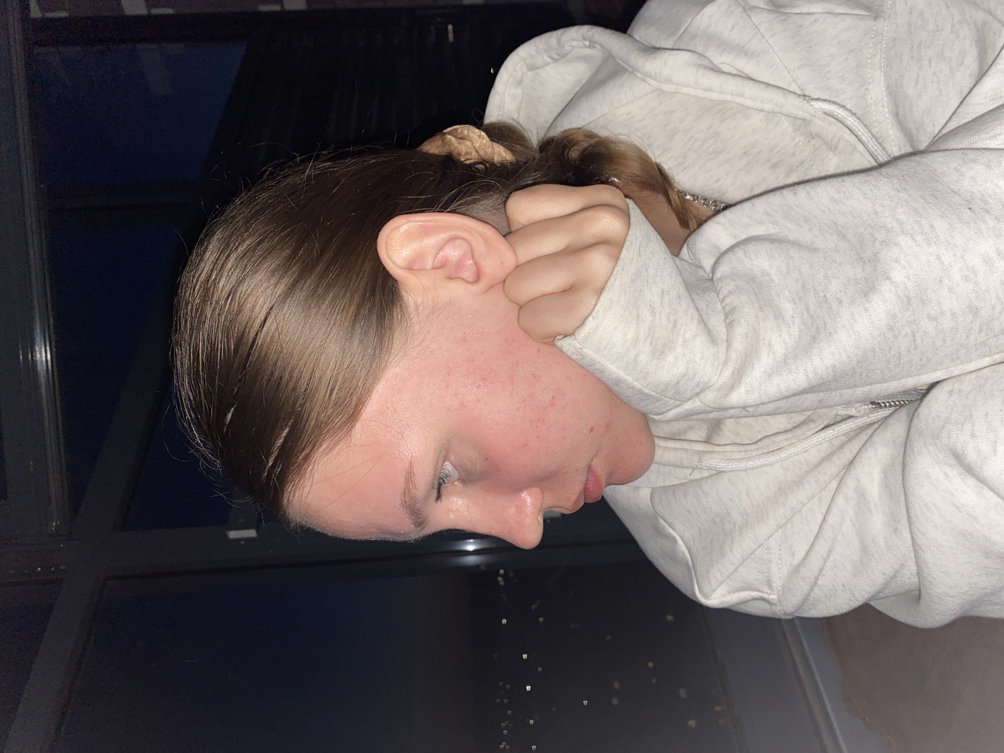 A close-up profile shot shows a young woman wearing a light gray hooded sweatshirt. Her left hand is raised, with her fingers touching her chin and cheek, partially obscuring her face. Her hair is pulled back into a ponytail with a scrunchie. She is looking to her left, out a large window. Outside the window, blurred lights of a city can be seen in the distance, suggesting an urban setting at night. The background within the room is dark, with a hint of what appears to be window framing. The image is taken indoors, likely in a high-rise building given the distant city lights. There is no identifiable information to suggest the location is Izhevsk, Russia.