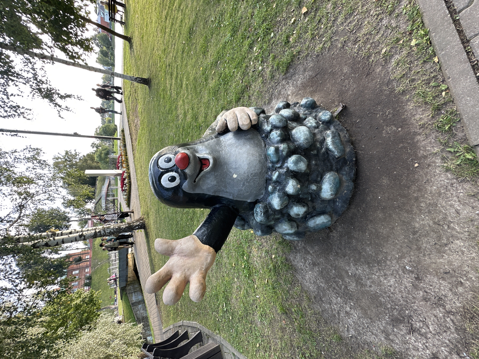 The image shows a statue of a cartoon mole, presumably "The Little Mole" character, standing in a park-like setting. The mole statue is grey with large black eyes and a red nose. It is emerging from a mound of dark, greenish-blue rocks. The mole's left hand is raised in a wave, with five fingers visible. Its right hand is resting on the rock mound. The statue is situated on a dirt patch surrounded by green grass.
In the background, there is a pathway made of paving stones, with people walking on it. A bridge with railings is visible in the mid-ground, spanning over a canal or body of water. Beyond the bridge, there are trees and a red brick building with windows. A body of water, likely a river or lake, is visible in the distance, with trees and buildings along its banks. The sky is bright and appears to be daytime.
The location is identified as Sergiyev Posad, Russia, and the statue is situated in a public park area, likely along the Dubna River, which flows through the city. The overall environment suggests a recreational area with a walking path, benches (one is visible on the far right), and manicured greenery. The presence of people suggests it is a popular spot for recreation.