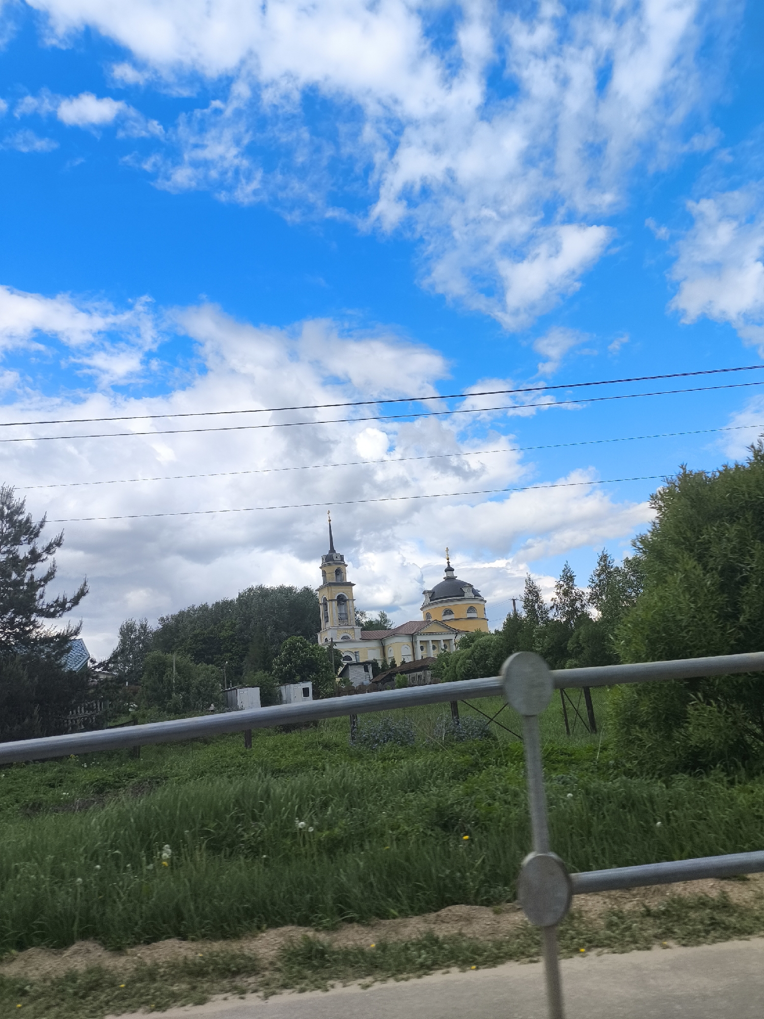 The image depicts a building, possibly a church, with a tall, yellow-toned tower and a rounded dome, partially obscured by trees. The sky is predominantly blue with patches of white clouds. A metal fence runs horizontally across the lower half of the image, with green vegetation in the foreground. The photograph appears to have been taken from a moving vehicle, based on the presence of the fence. The location of the building is in an unknown city, unknown.