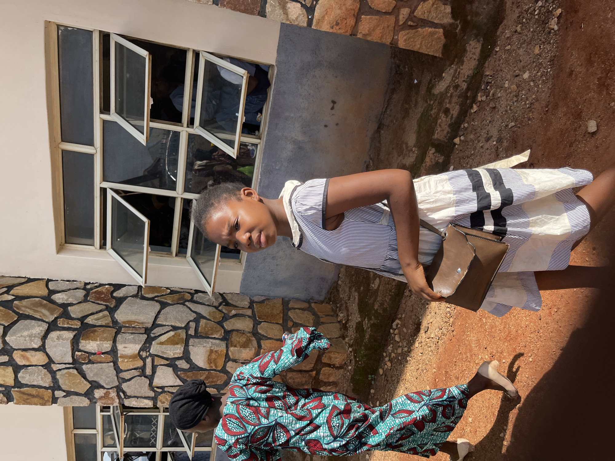 A woman in a striped dress and shoulder bag stands next to another woman in a colorful patterned outfit. They are outside a building with stone walls and open windows. The location is Gura Top, Nigeria.
