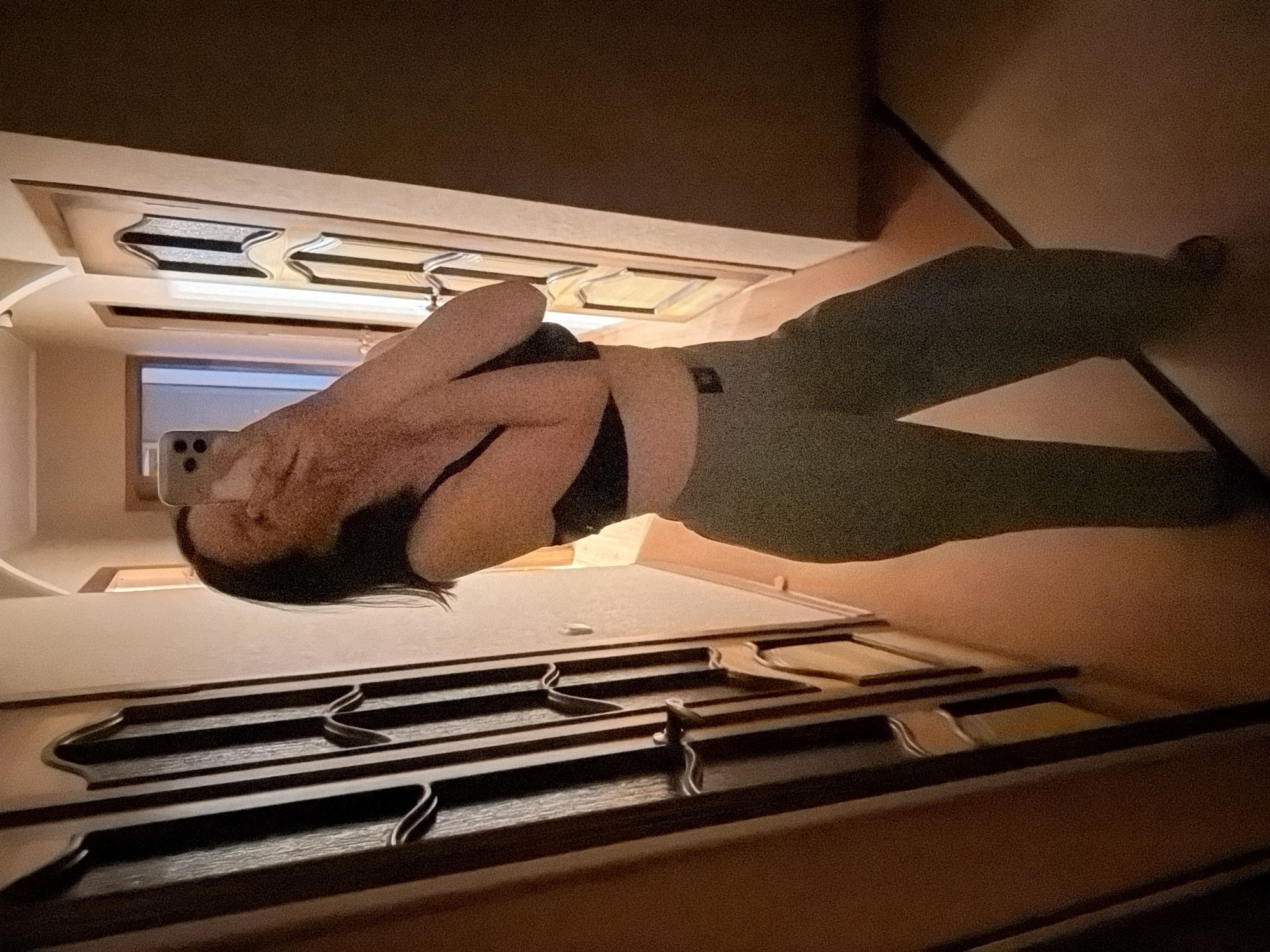 A person is posing in what appears to be an interior setting, possibly a home. The individual is holding a smartphone, potentially taking a selfie. The person has dark hair and is wearing a dark-colored top with light-colored pants. The background includes architectural details such as wooden furniture or cabinetry, and ceiling fixtures with ornate designs. Outside a window, there is a glimpse of daylight. The specific location within a city is Unknown, Unknown.