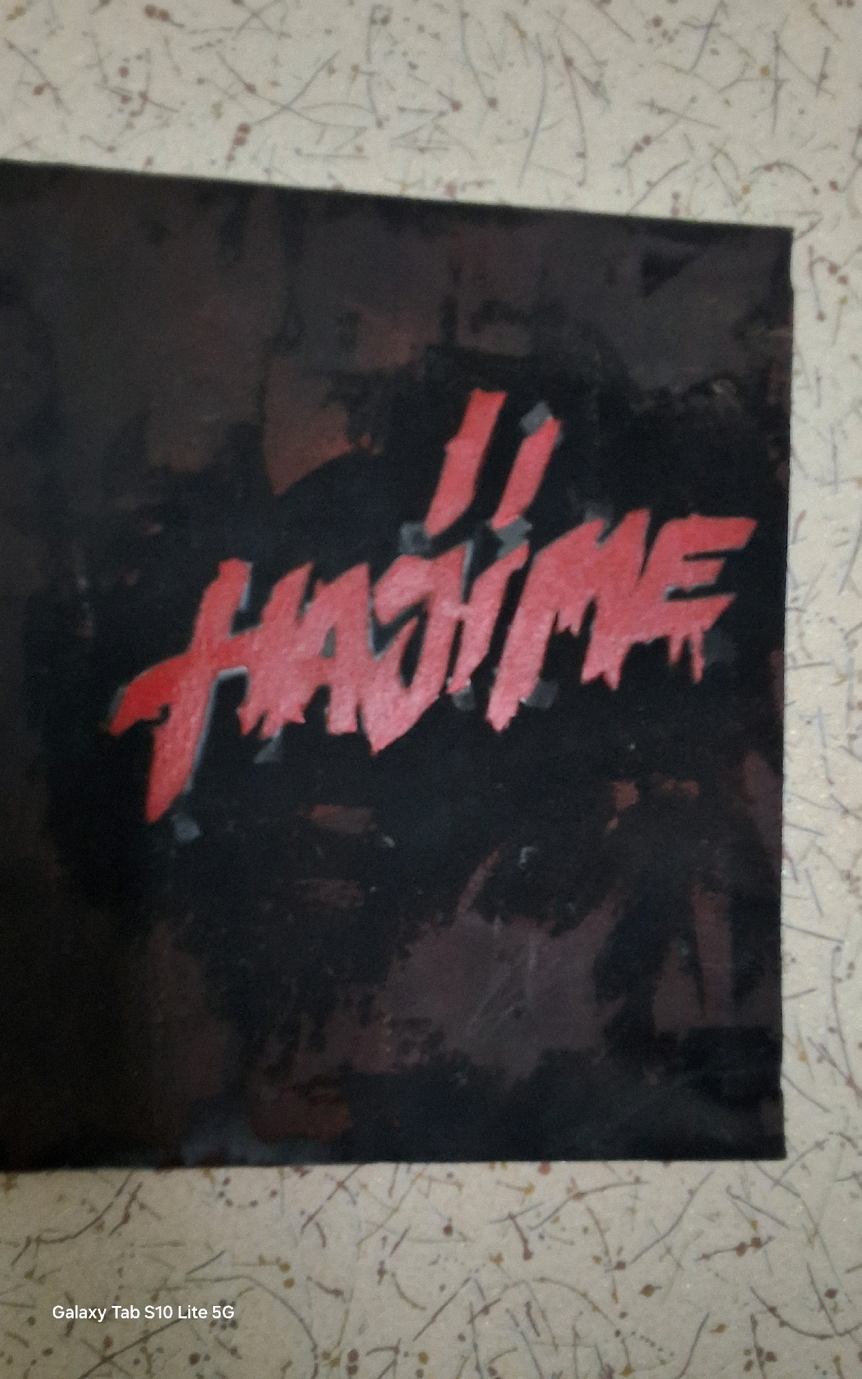 The image is a close-up, slightly angled photograph of a black canvas with red lettering that reads "JI HAJIME." The lettering appears stylized and somewhat distressed, with a ragged, dripping effect. The background of the canvas is black, with some subtle texture or smudges that add to a dark, moody aesthetic. The canvas is placed against a light-colored wall with a speckled pattern, possibly wallpaper, that has thin, irregular lines and dots in shades of brown and red. A watermark at the bottom left corner of the image indicates "Galaxy Tab S10 Lite 5G." There is no indication of the city of Gal, Abkhazia, or any specific geographical location. The image does not contain any people, actions, or interactions.