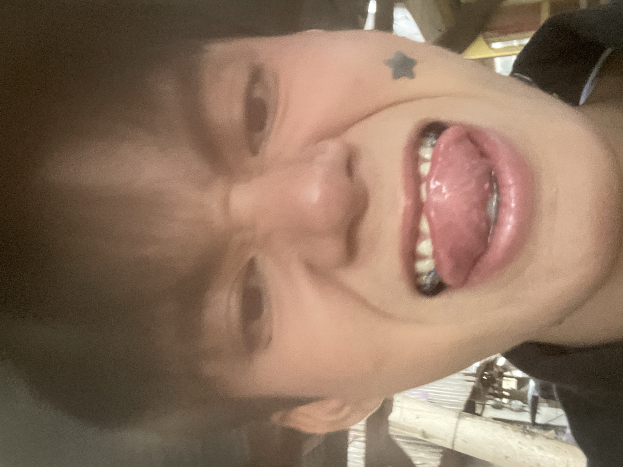 The image shows a close-up, low-angle selfie of a young person with their tongue sticking out. The individual has dark hair, braces, and a small star tattoo on their cheek. They are wearing a dark shirt. The background appears to be outdoors, with some corrugated metal and wooden structures visible, suggesting a simple or possibly rural setting. There are no clear indicators in the image that would allow for a specific location within Davao City, Philippines to be identified.