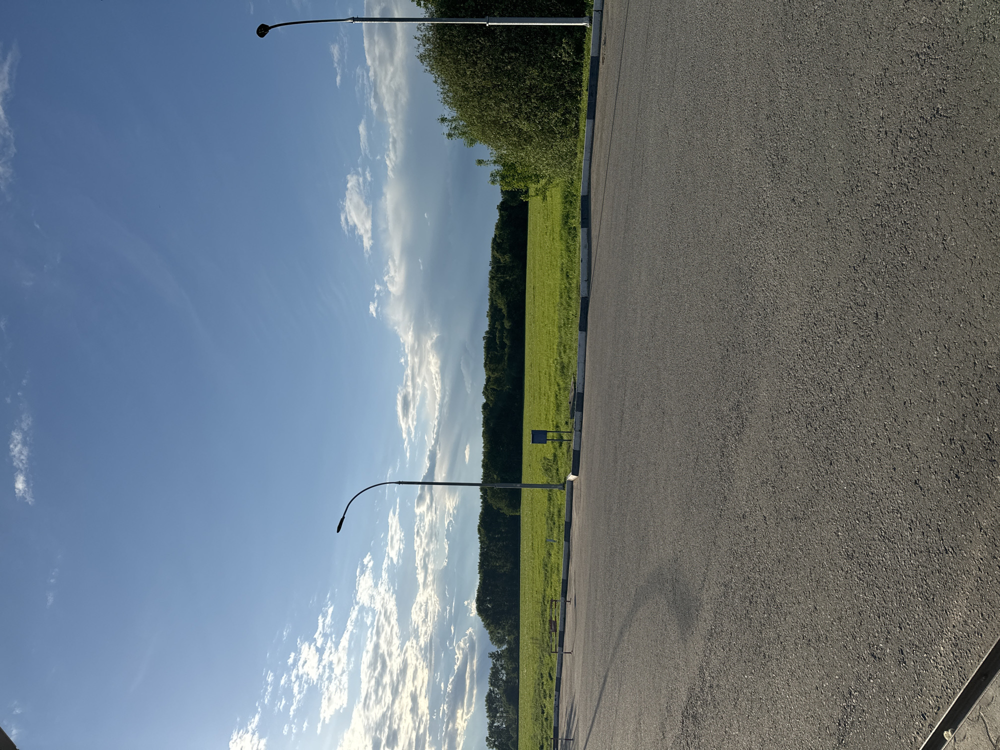 The image shows a section of asphalt road in Bolshiye Vyazyomy, Russia. The road is bordered by a curb and a grassy area with trees. The sky is blue with scattered clouds. Two streetlights are visible along the roadside. There are no people or other subjects in the image.