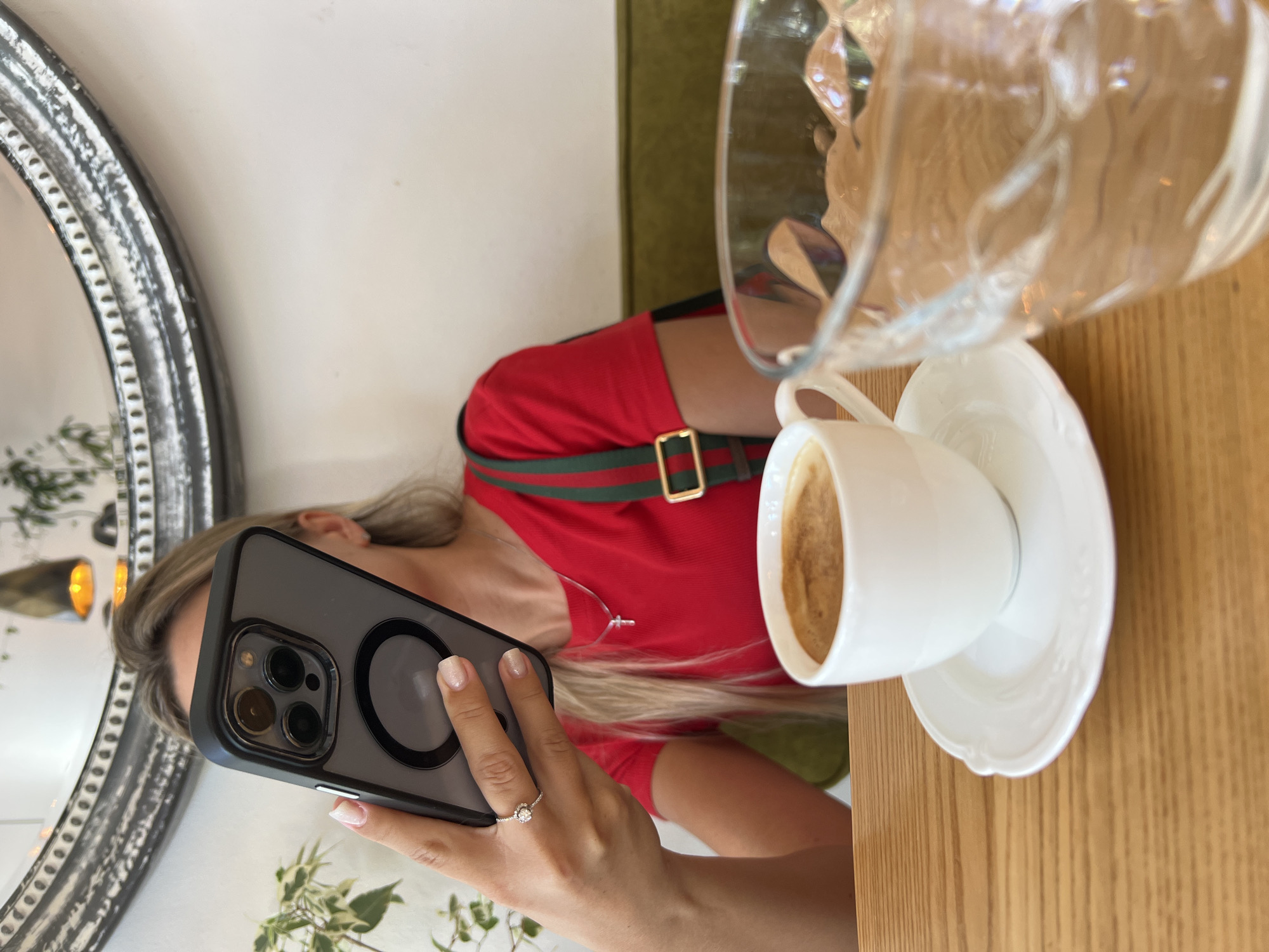 A person is sitting at a wooden table in what appears to be a cafe in Odesa, Ukraine, holding a smartphone in their right hand. The person is wearing a red t-shirt with a green and red strap across their shoulder. In front of them, on the table, is a white cup of coffee and a partially filled glass of water. The background shows a decorative mirror and a hint of green upholstery. The lighting suggests it's daytime.